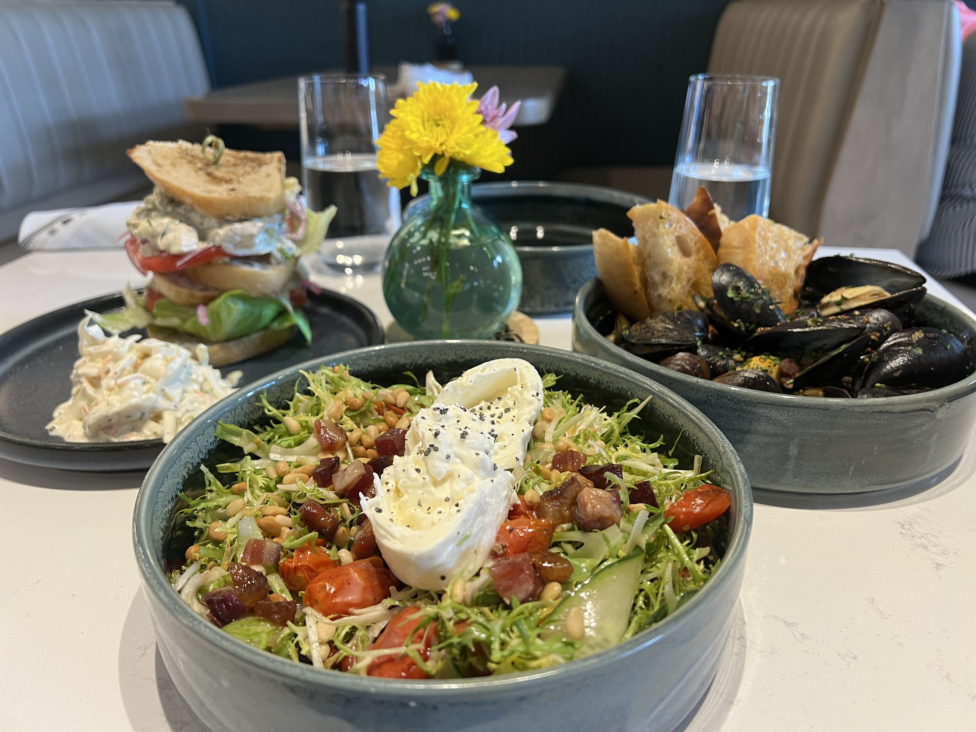 Table with a large greens salad topped with burrata; a mussel and bread dish; a stacked sandwich; a creamy slaw on the side; yellow flowers in a teal vase in the background; two water glasses.