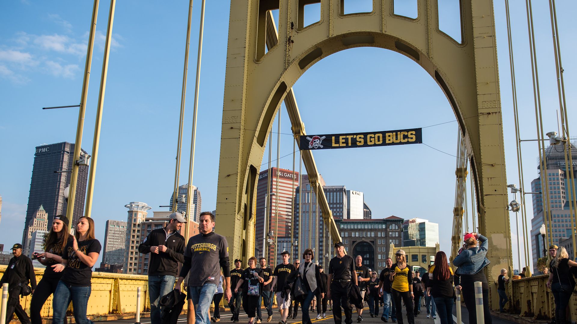 Crowd wearing black and yellow Pittsburgh Pirates apparel walking on a yellow steel bridge under a banner that reads "LET'S GO BUCS" with city skyscrapers in the background.