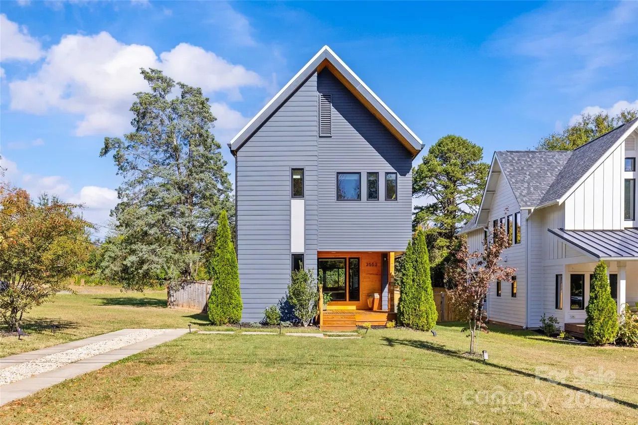 Modern two-story gray house with triangular roof and wooden porch, surrounded by green grass, trees, and neighboring white house under a blue sky with clouds.