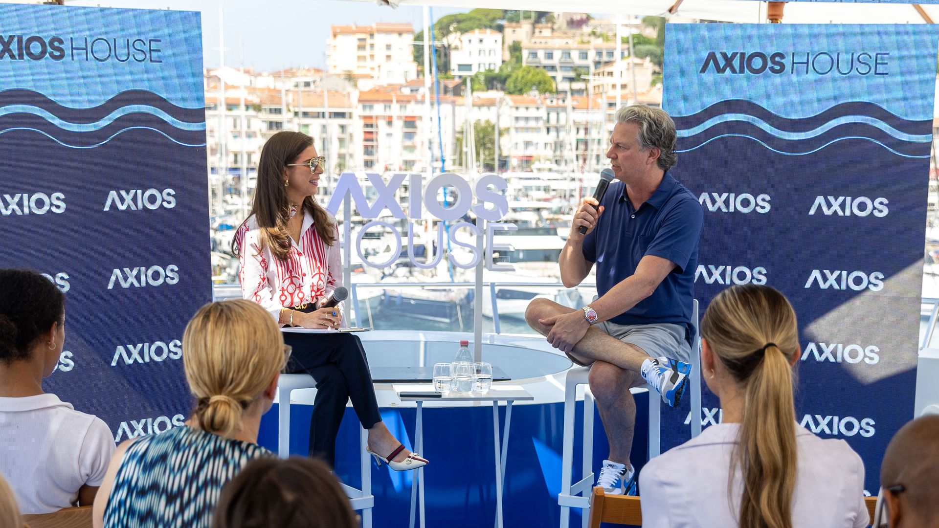 Outdoor interview on a boat: Axios' Sara Fischer and People Inc. CEO Neil Vogel with microphones talk on a blue Axios House stage, flanked by banners. Audience sits, marina and buildings visible in the background.