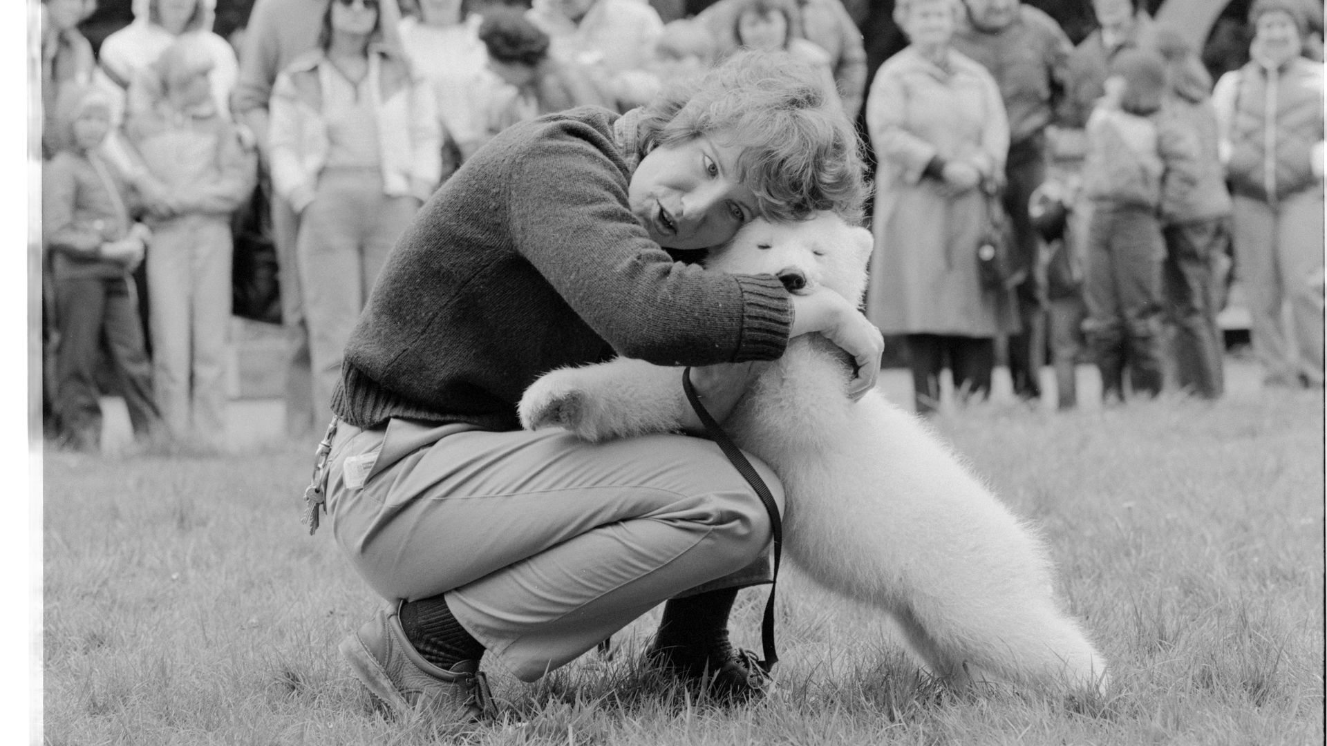 Black-and-white photo of a veterinarian hugging a small white baby polar bear as a crowd looks on