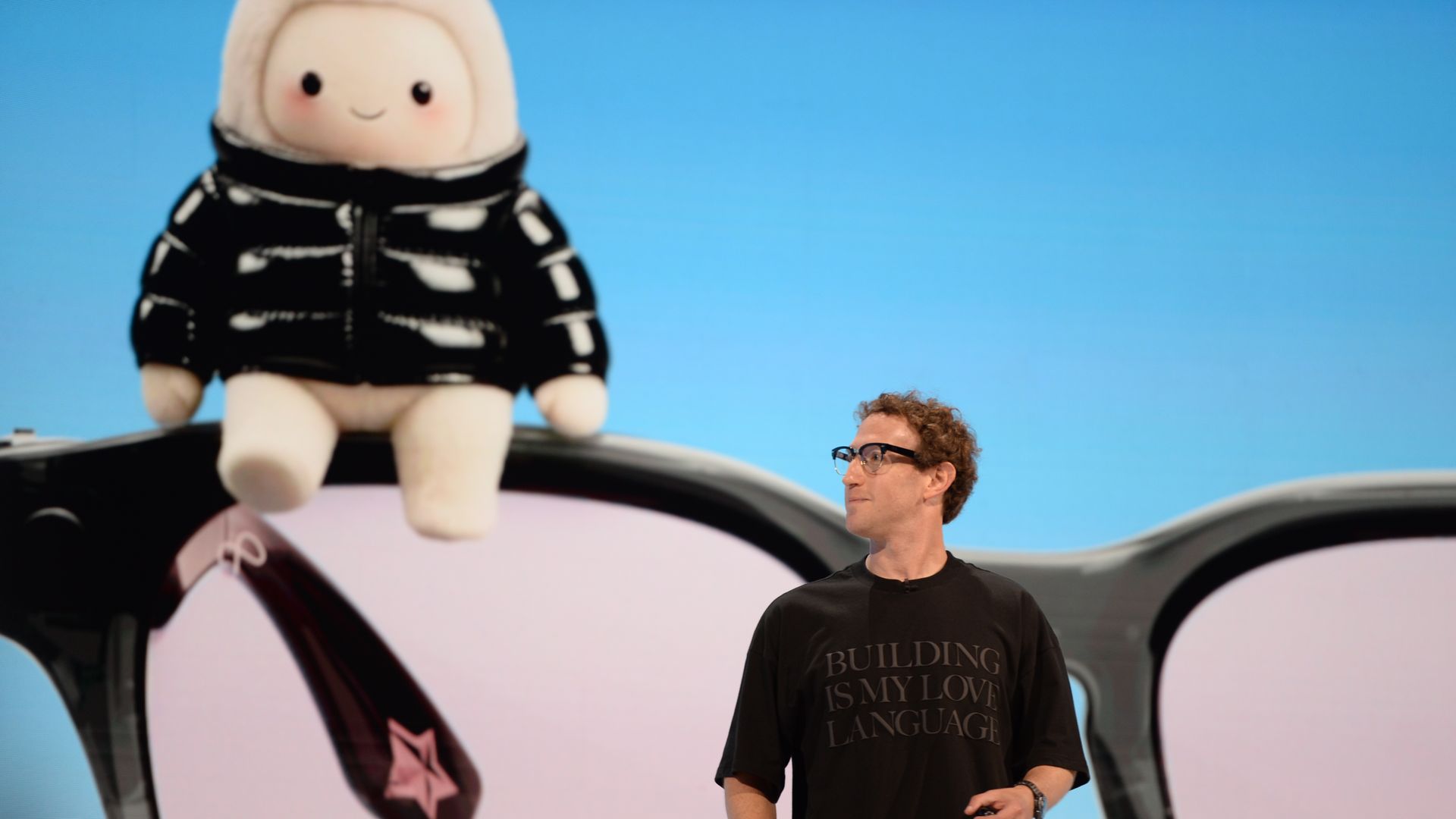 A man with glasses wearing a black shirt that reads "BUILDING IS MY LANGUAGE" stands beside a large white plush doll in a black-and-white striped hoodie perched on a car, with a bright blue sky behind.