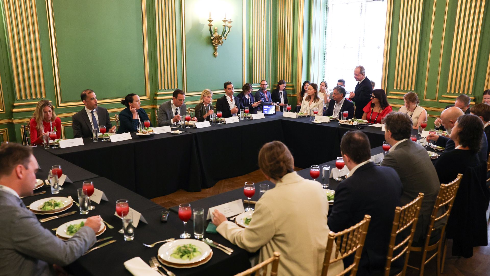 Group of professionals in formal attire seated around a U-shaped table with black tablecloths in an ornate green room during a roundtable discussion.