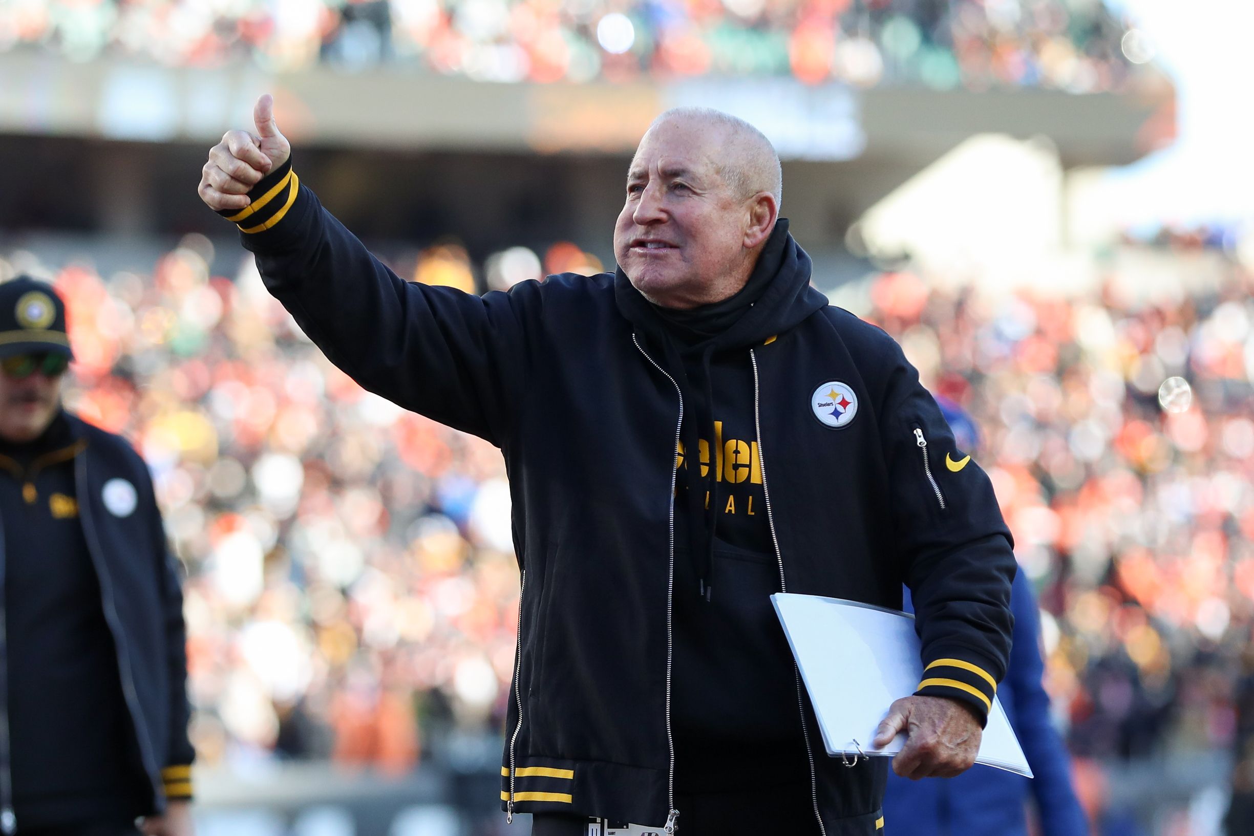 An older man with a gray buzz cut wears a black and gold sweat suit and t-shirt. A large wad of gum is visible in his cheek. 