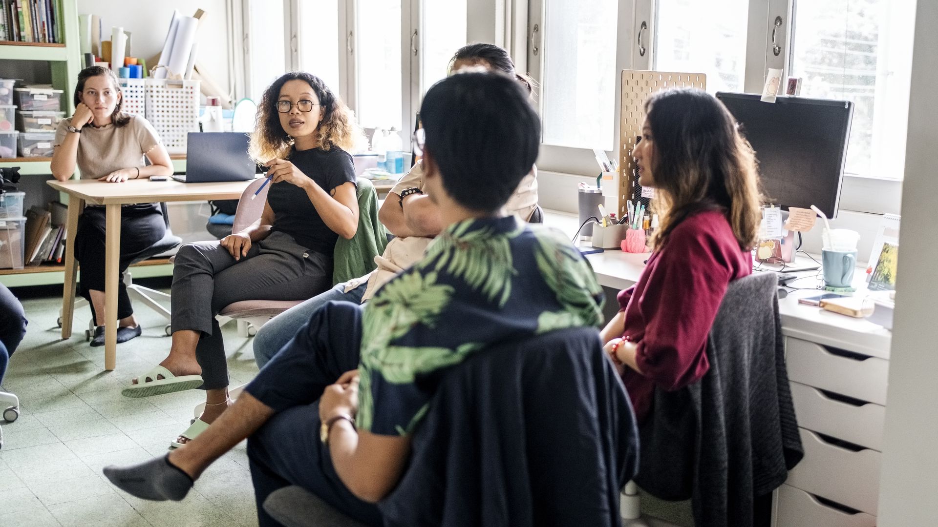 A diverse group of five people engaged in a discussion in a bright room with large windows, desks and office supplies. One woman gestures while others listen attentively.