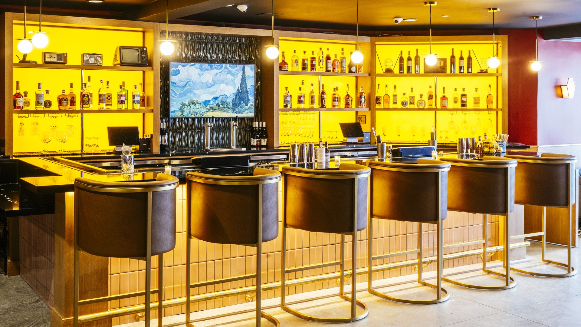 A modern-designed bar with six brass and leather bar stools lined up at it. The back-bar glows with amber lighting, and bottles are lined up along the back wall.