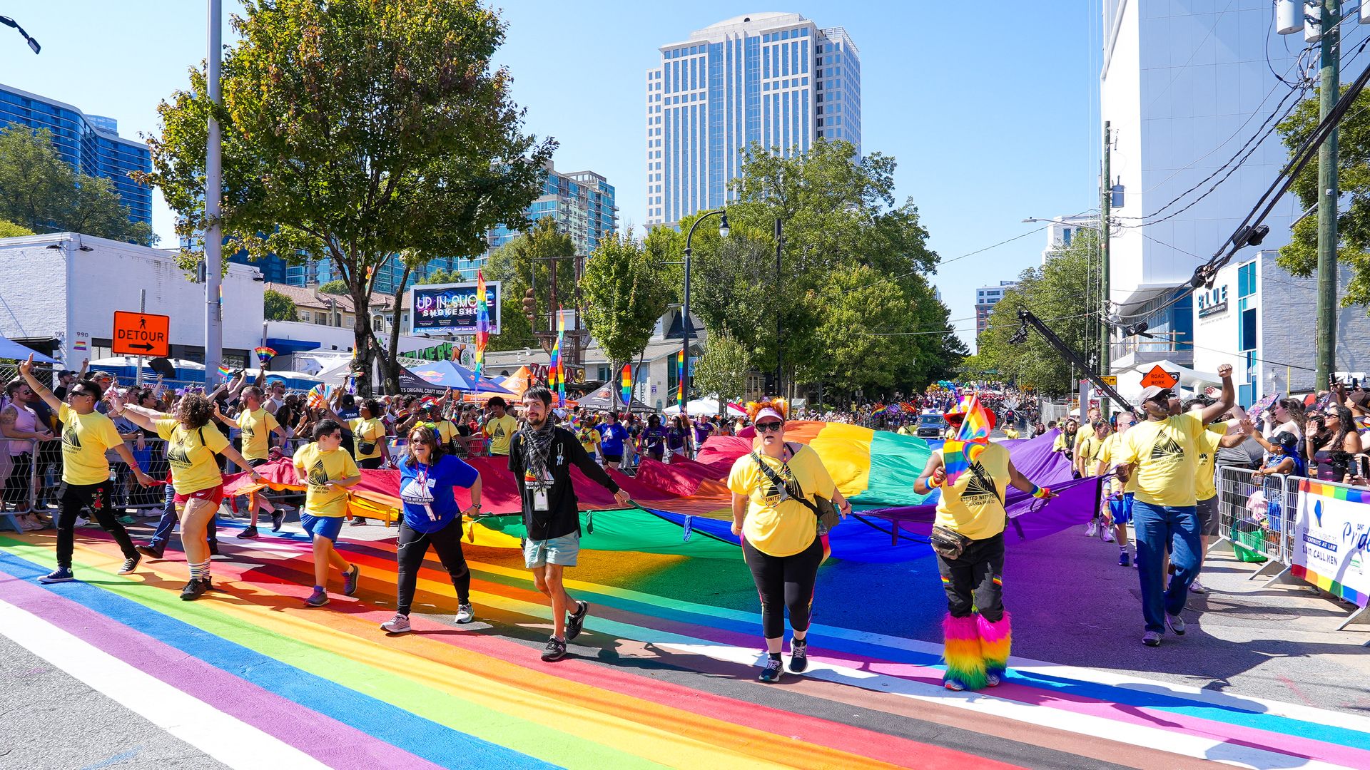 People wearing yellow shirts and carrying a super-sized rainbow flag in an urban area with rainbow crosswalks on a sunny day.