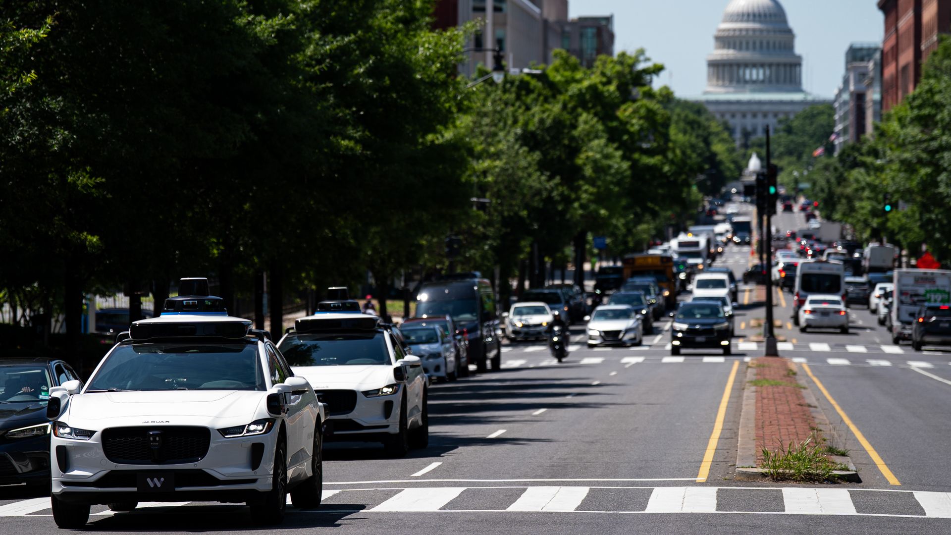 Two of Waymo's test vehicles drive down D.C. street with the U.S. Capitol in the background.