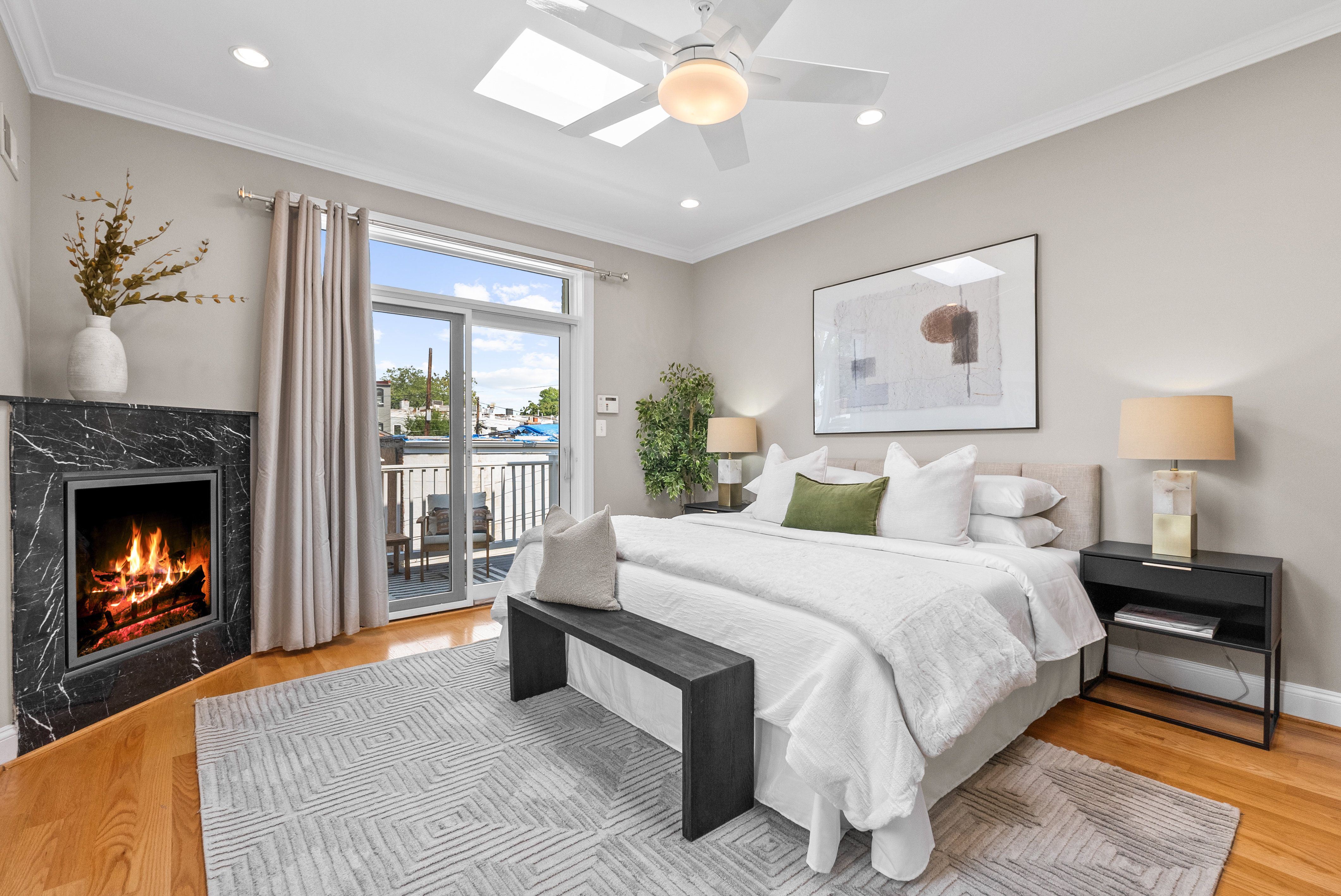 Bright bedroom with a white bed, green decorative pillow, black bench, gray geometric rug, black marble fireplace with fire, beige walls, two lamps, a plant, and sliding glass doors to a balcony.