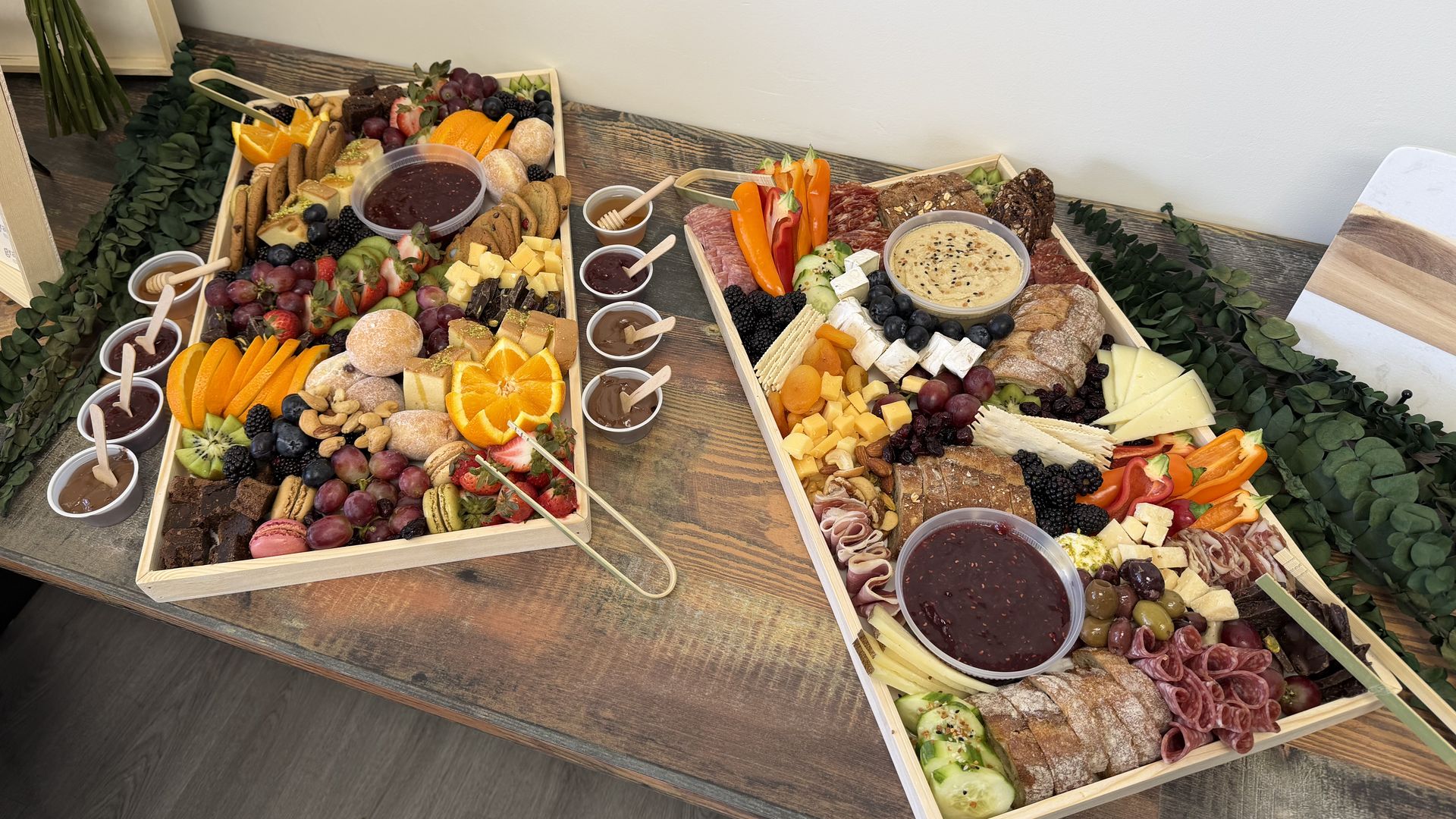 Two wooden charcuterie boards on a rustic table, piled with cheeses, cured meats, crackers, bread, fruits (grapes, berries, orange slices, kiwi), nuts, and two bowls of dark dipping sauce.