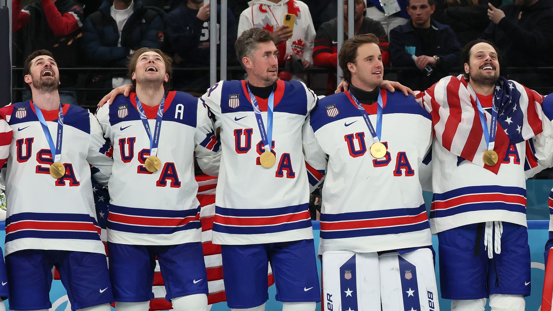 USA men's ice hockey team in white jerseys with red and blue stripes celebrating with gold medals, one player wrapped in American flag, standing on ice rink with fans behind them.