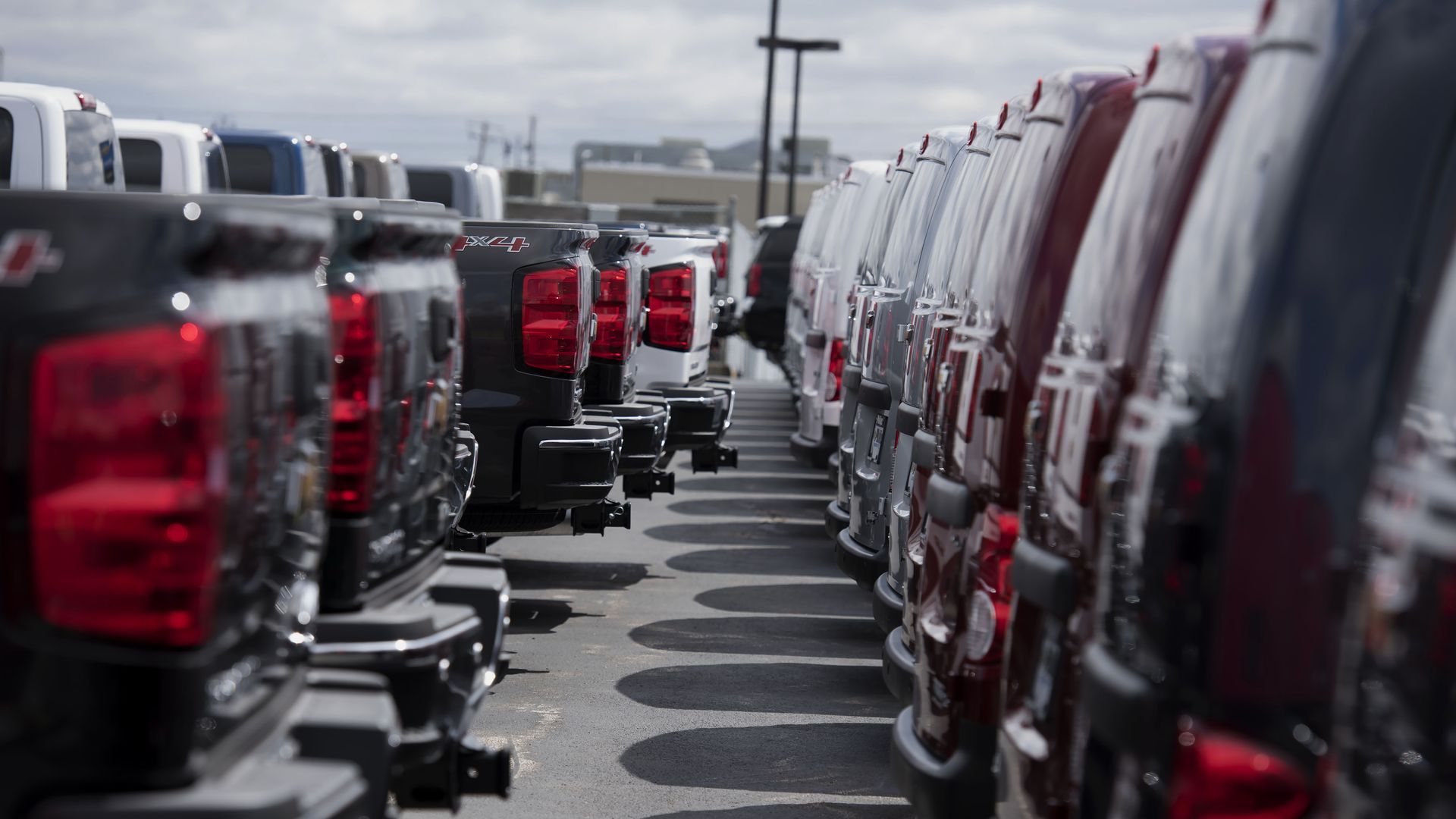Line of new pickup trucks parked bumper-to-bumper in a dealership lot, red taillights visible, black, white, and silver vehicles forming a narrow path between rows under a cloudy sky.