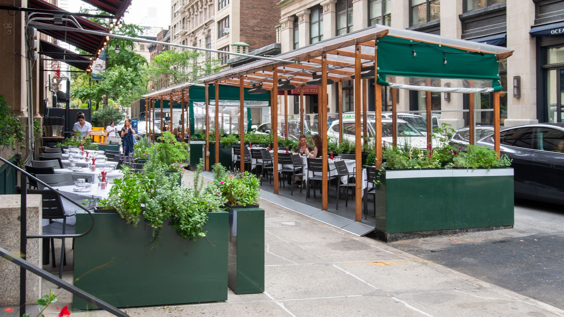 Urban sidewalk cafe with a row of white linen tables and black chairs under red striped awnings. Green planters border the dining area; wooden pergola with string lights and tall buildings nearby.