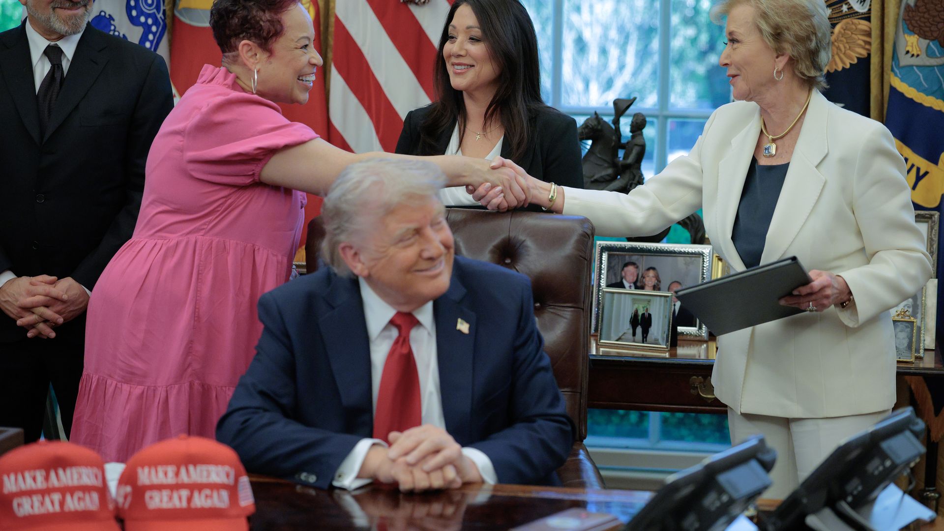 Man in navy suit and red tie sitting at desk with two red "Make America Great Again" hats, two women shaking hands—one in pink dress, the other in white pantsuit—with flags in background.