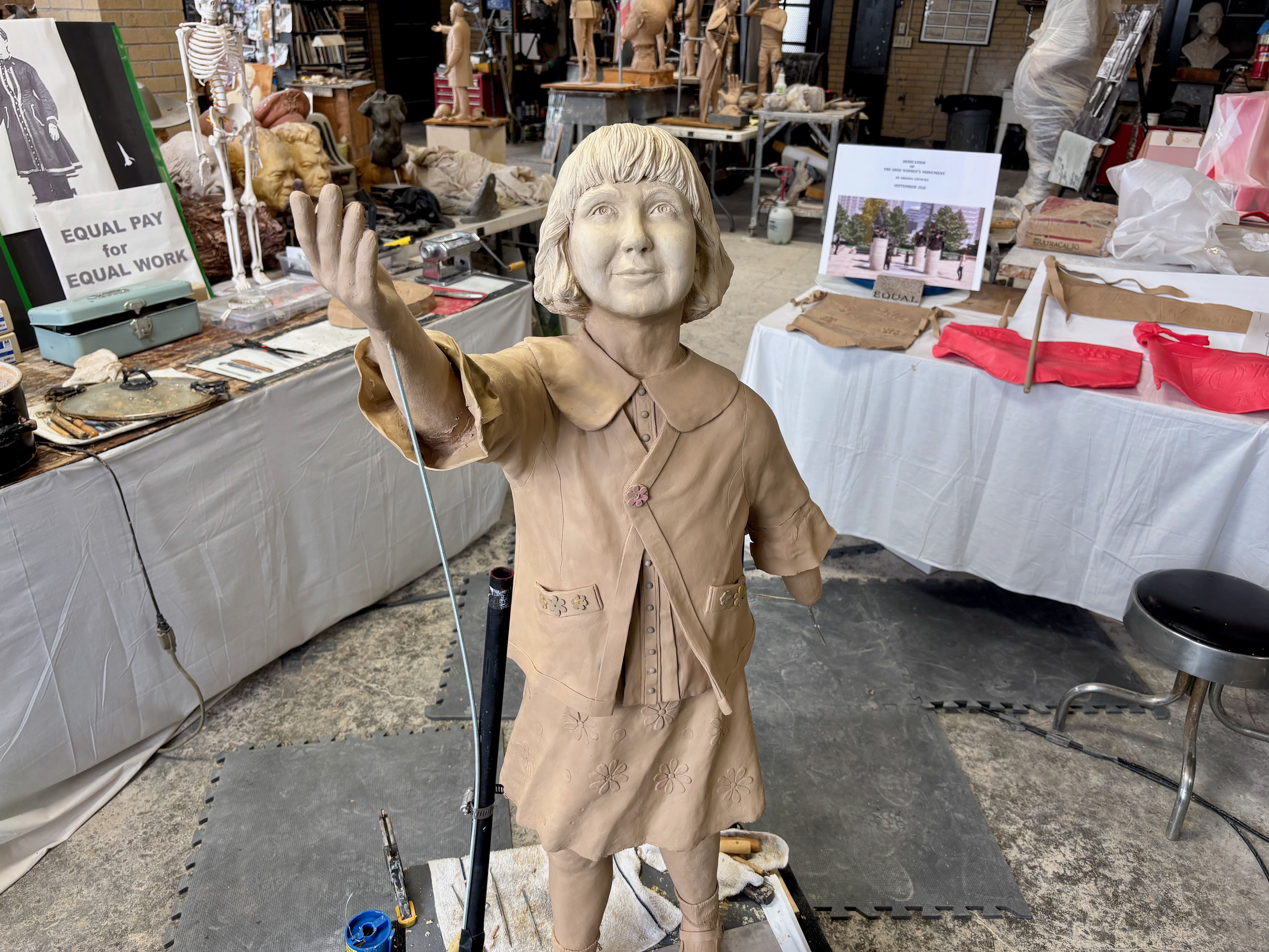 Life-size clay sculpture of a smiling girl with short hair in a beige dress, raising her right hand with a rod; art studio cluttered with sculptures, busts and supplies.