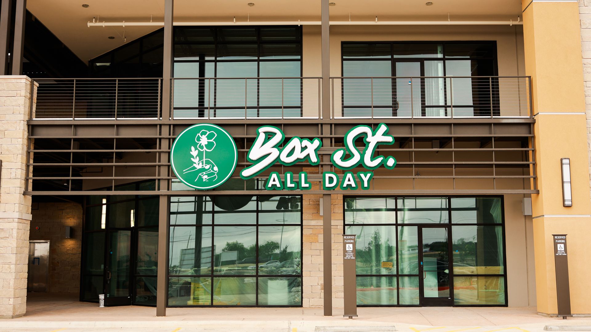 Modern storefront with large glass windows and a green circular logo reading "Box St. ALL DAY" on a beige building. Foreground shows a parking lot with a blue wheelchair symbol.
