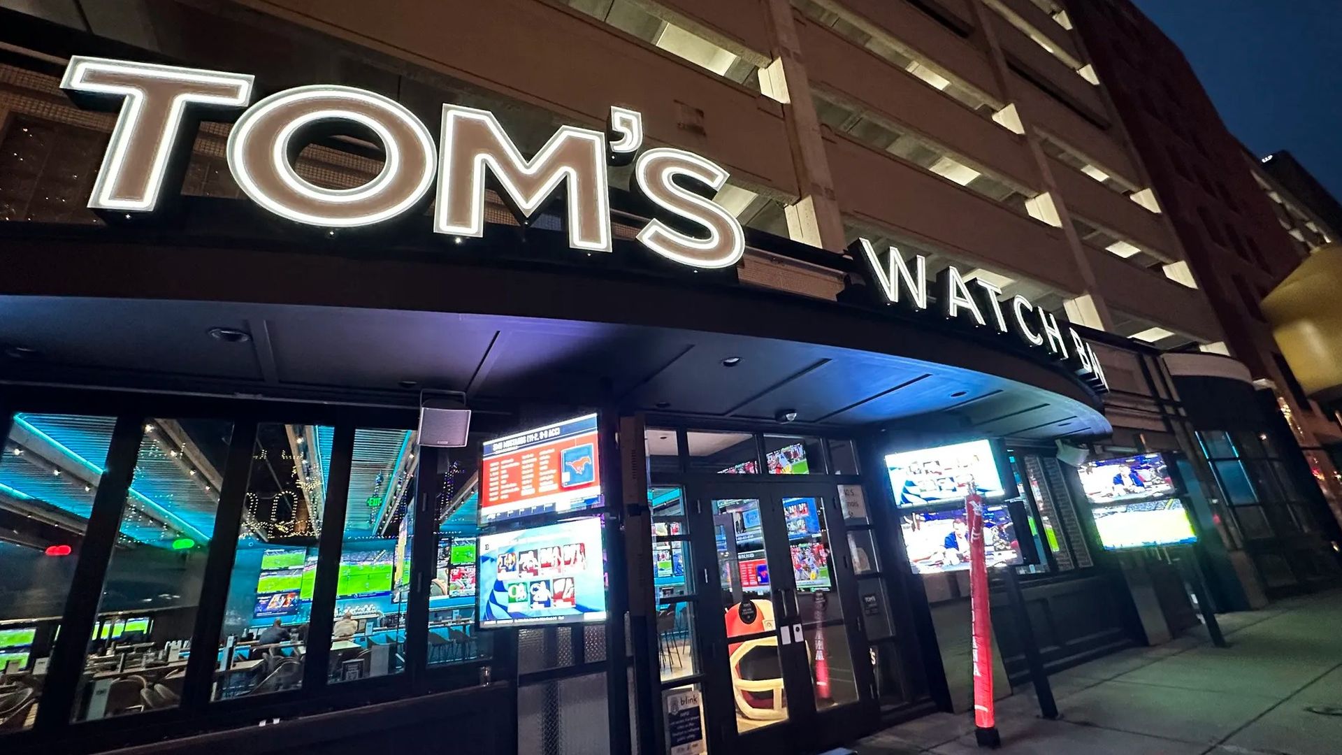 Night view of Tom's Watch Bar with bright white signage, illuminated interior featuring multiple TV screens showing sports, and neon blue interior lighting.