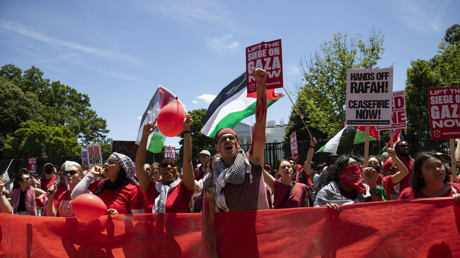 Pro-Palestinian protesters gather outside of the White House, and stand behind a large red banner, waving flags and holding protest signs.