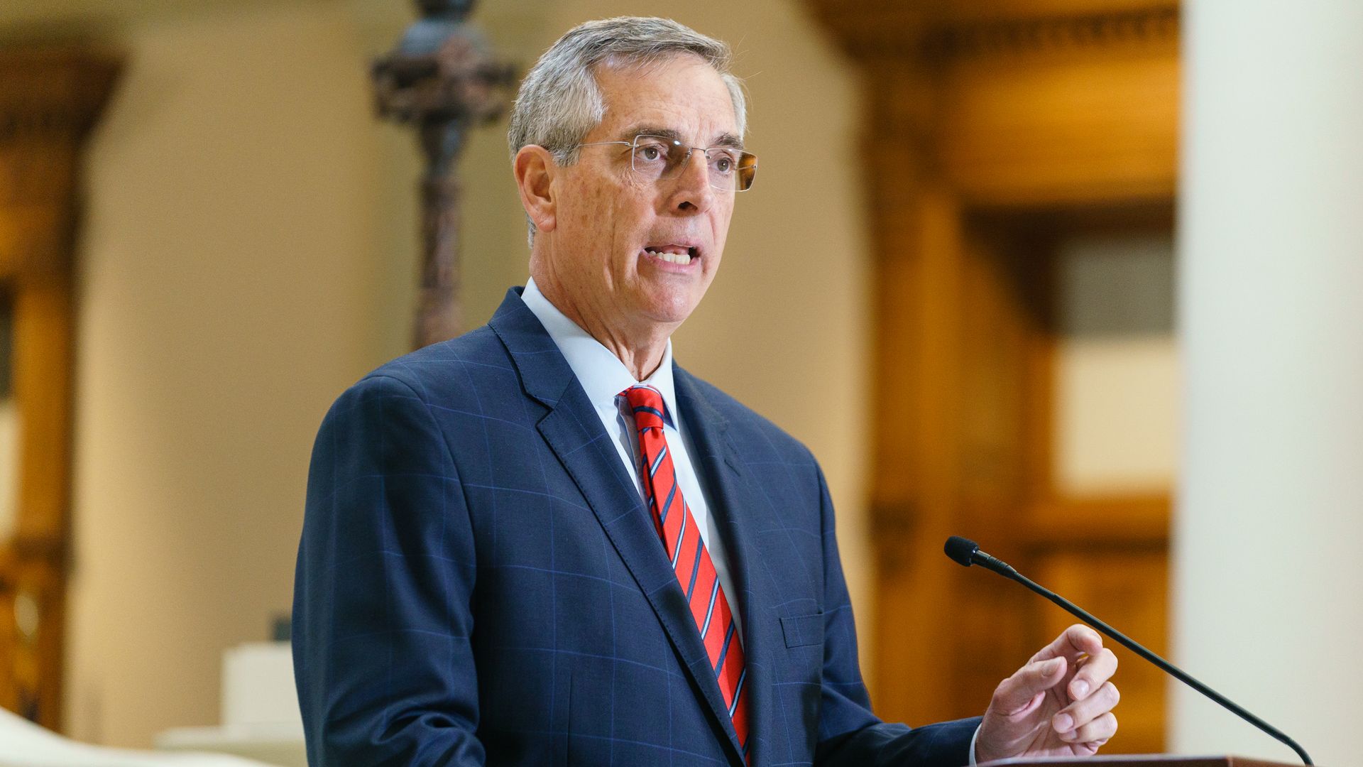 A man wearing eye glasses, a red tie with blue and white stripes and a blue suit speaks at a microphone.