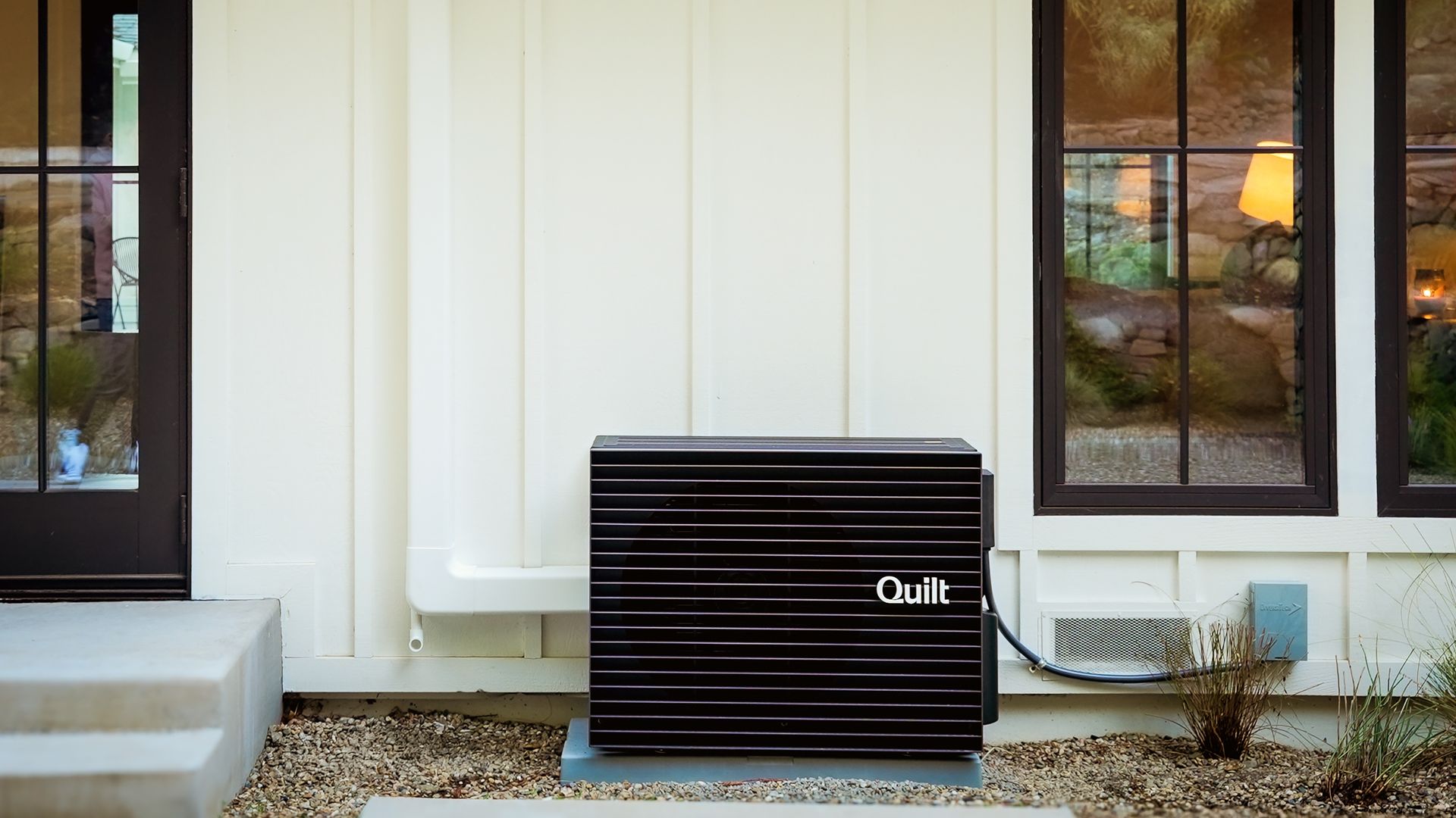 Black Quilt heat pump unit installed outside a white paneled house, near a black-framed glass door and window, on a gravel bed with some plants nearby.