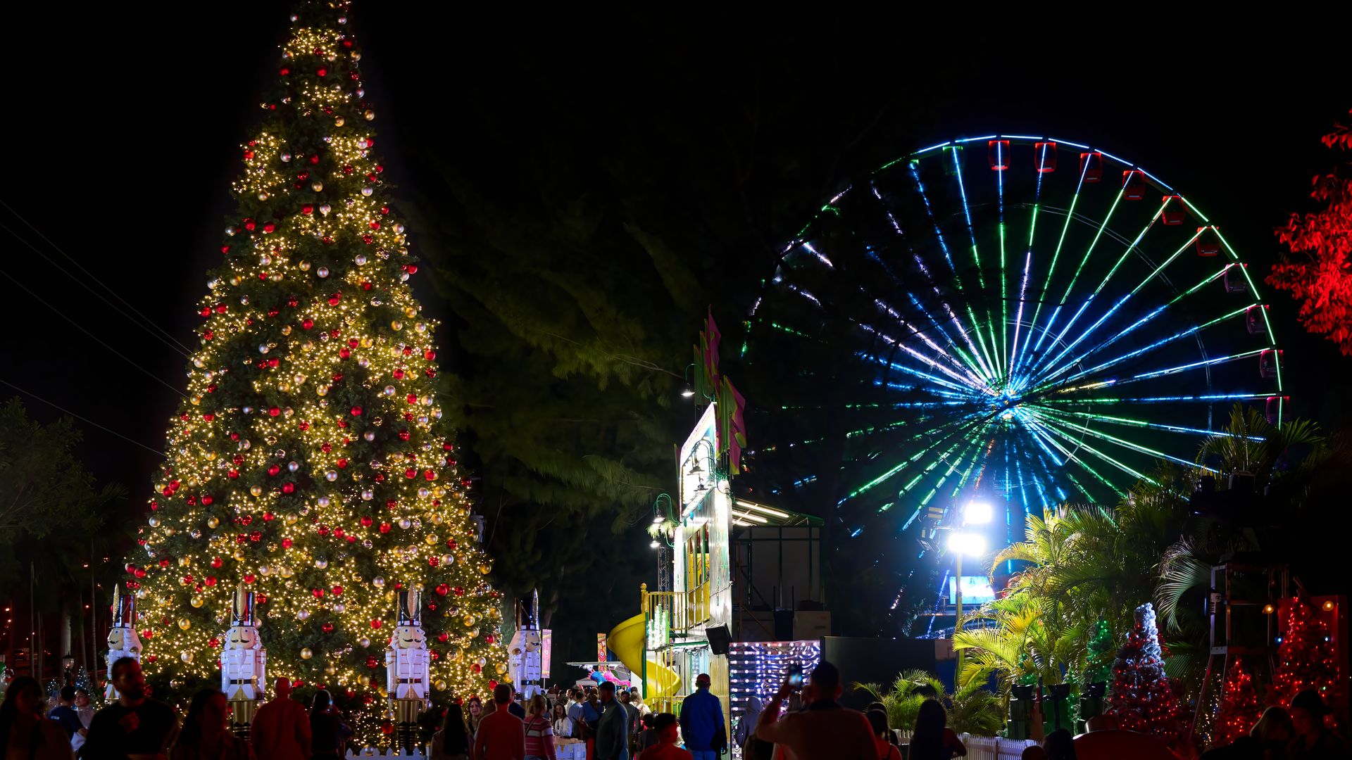 Night scene at a Christmas festival with a large decorated tree, illuminated nutcracker figures, a lit Ferris wheel in blue and green, and people walking and enjoying the event.