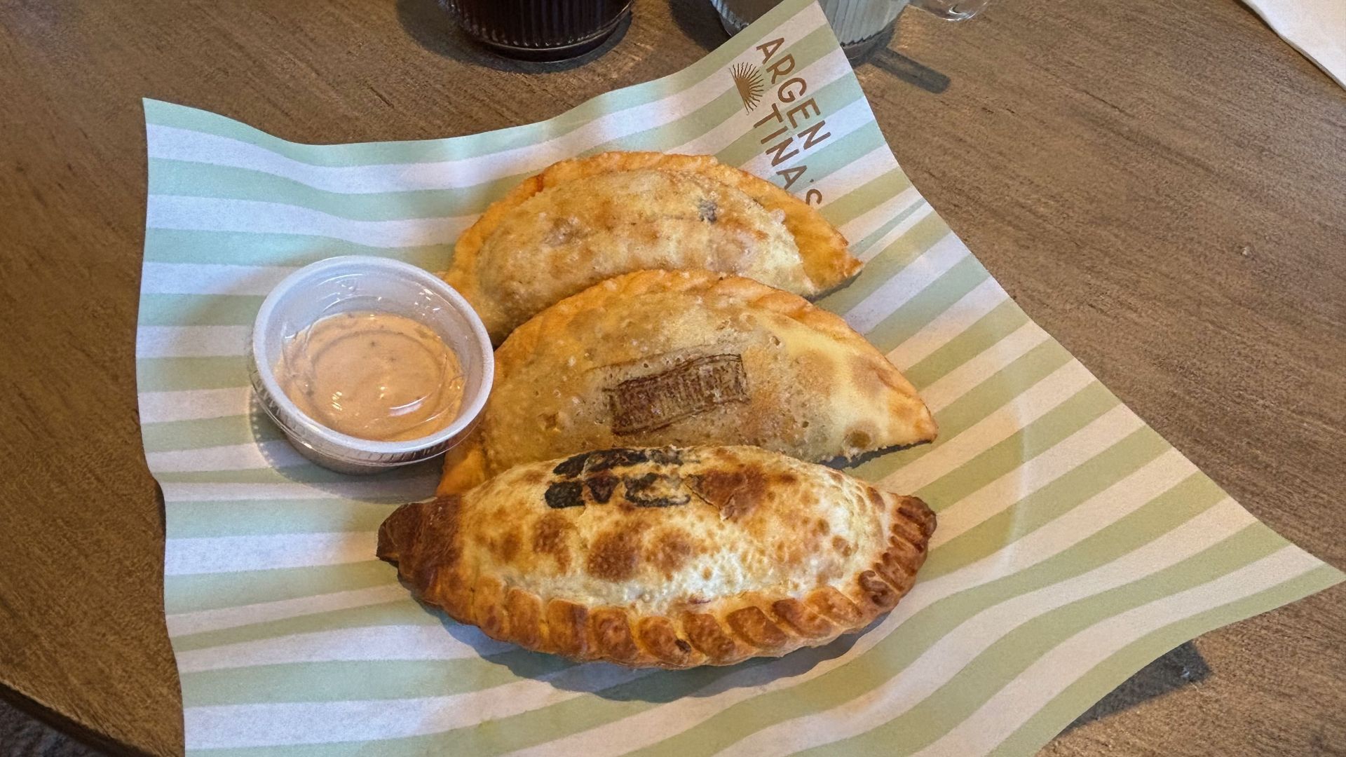 Three golden-brown empanadas on a green striped paper liner with a small cup of creamy dipping sauce, set on a wooden table.