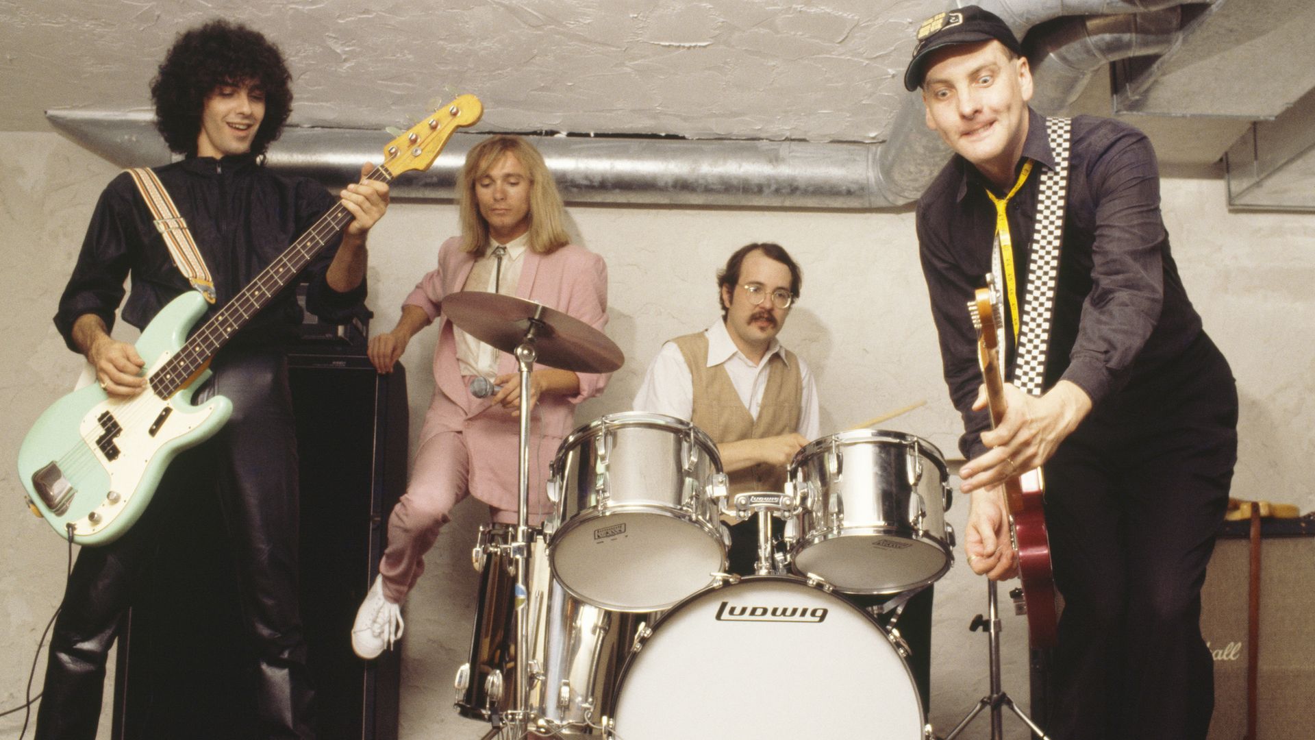 Man with guitar, man in pink suit, man sitting behind drums, man with guitar.