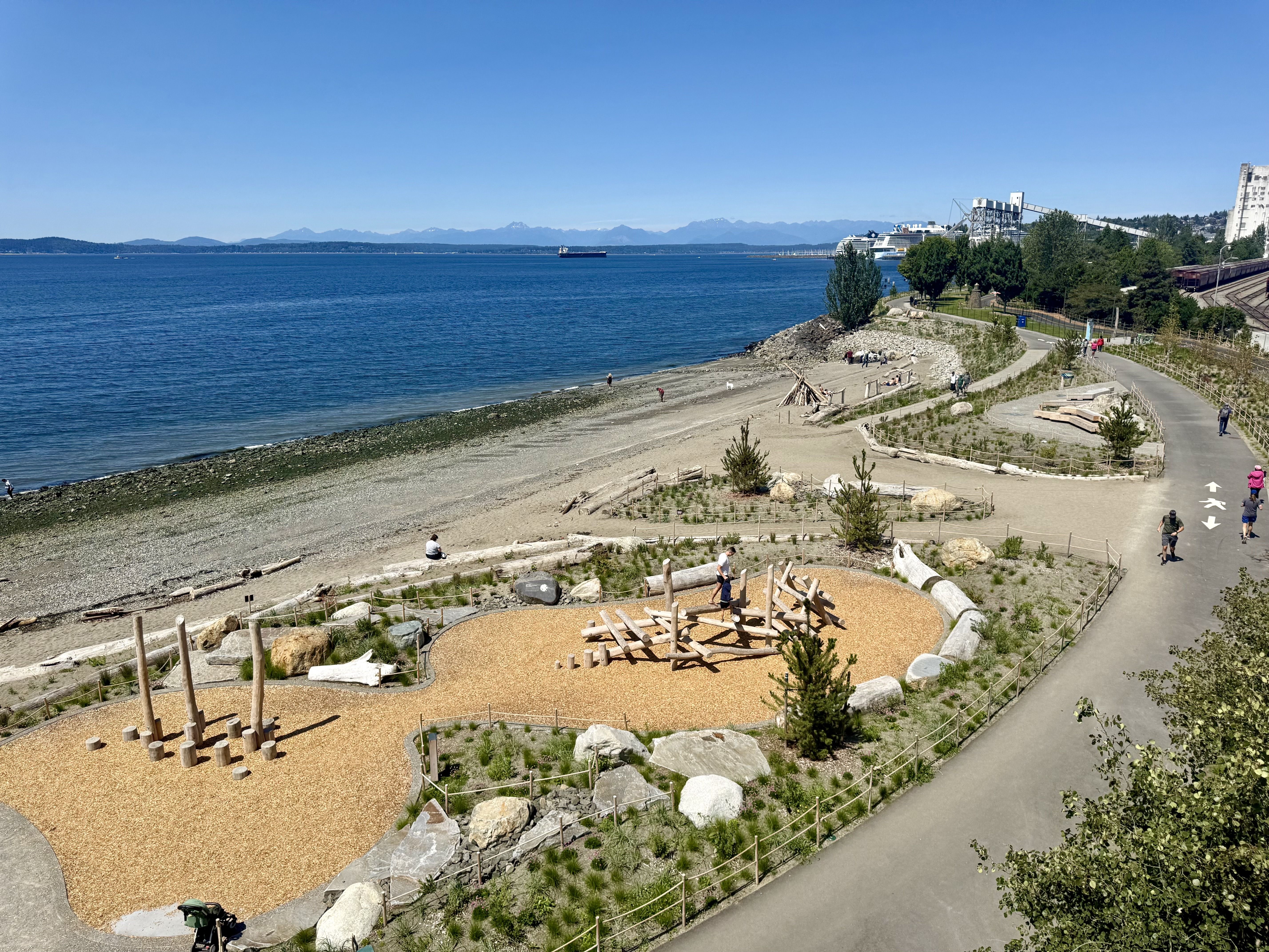 Wide coastal park along a blue bay with a curved path. Beach with wooden posts in the foreground, people walking, a white cruise ship and cranes to the right, trees and distant mountains.