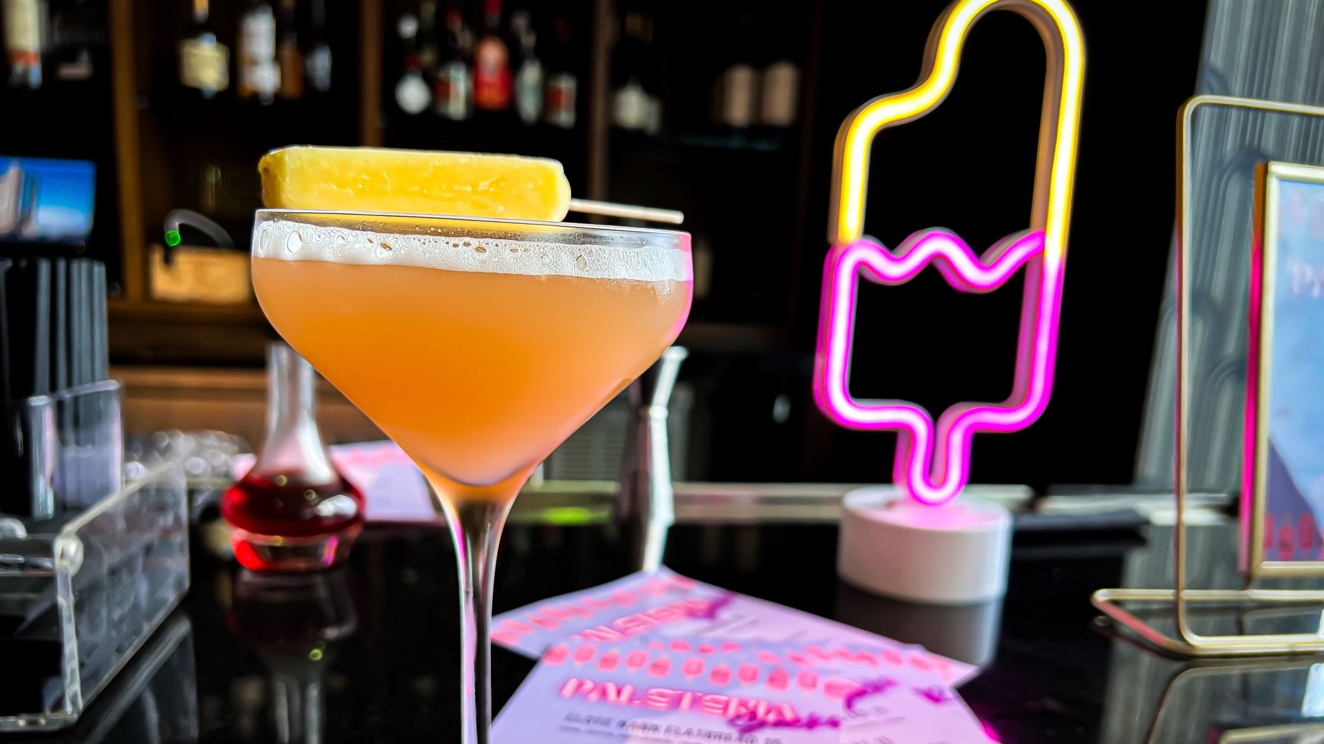 An orange-colored cocktail in a coupe glass topped with a small yellow paleta. A neon paleta sign is in the background.