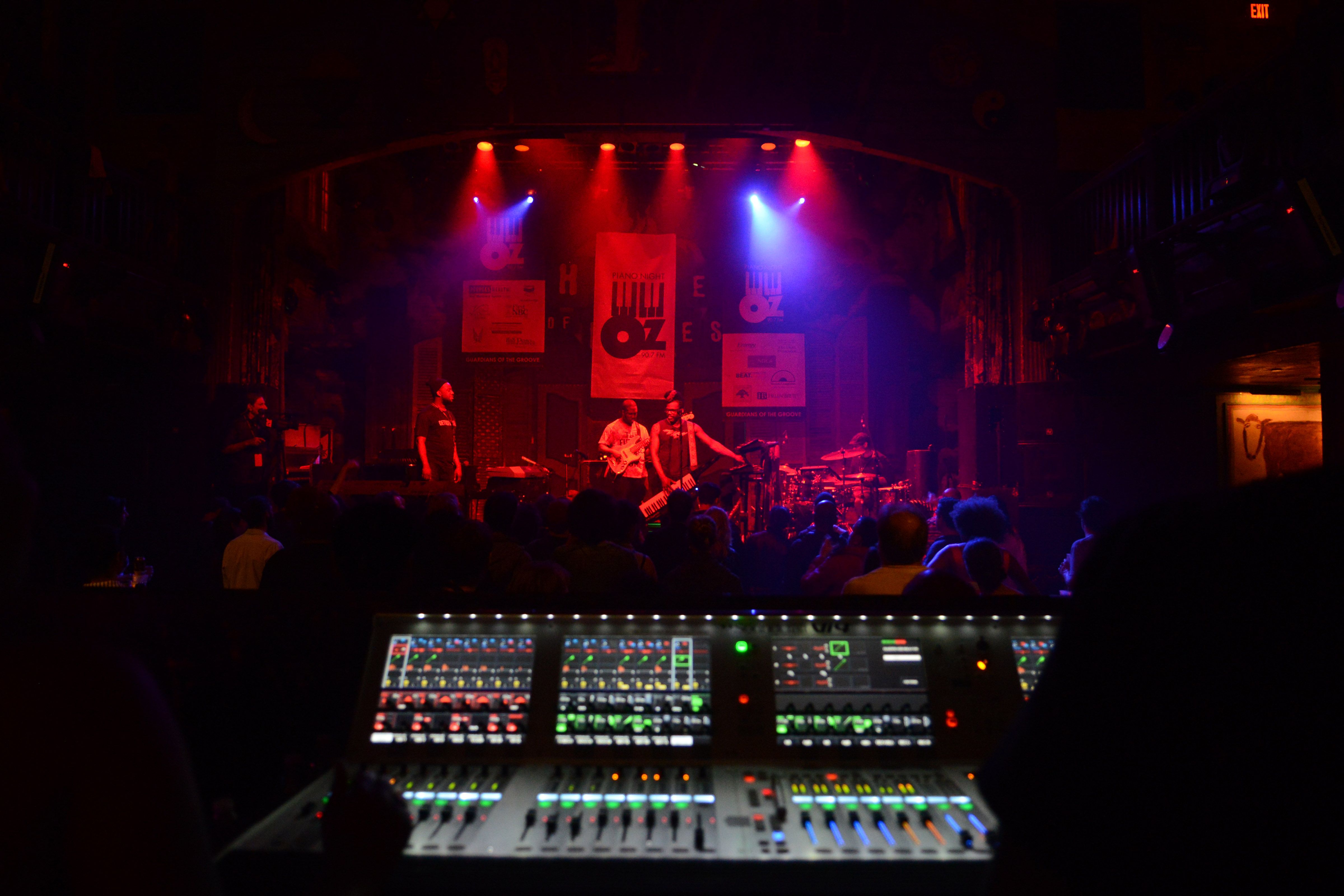In the background, Robert Glasper performs with his band onstage. In the foreground, sound board controls are lit up in a dark venue.