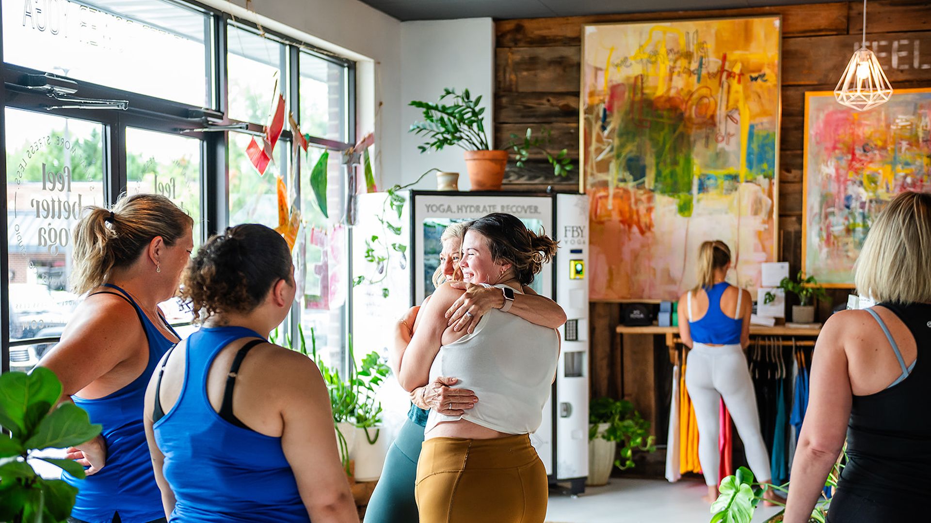 Group of women in colorful workout clothes hugging and chatting inside a bright yoga studio with plants, art, and a dog on a woven rug.