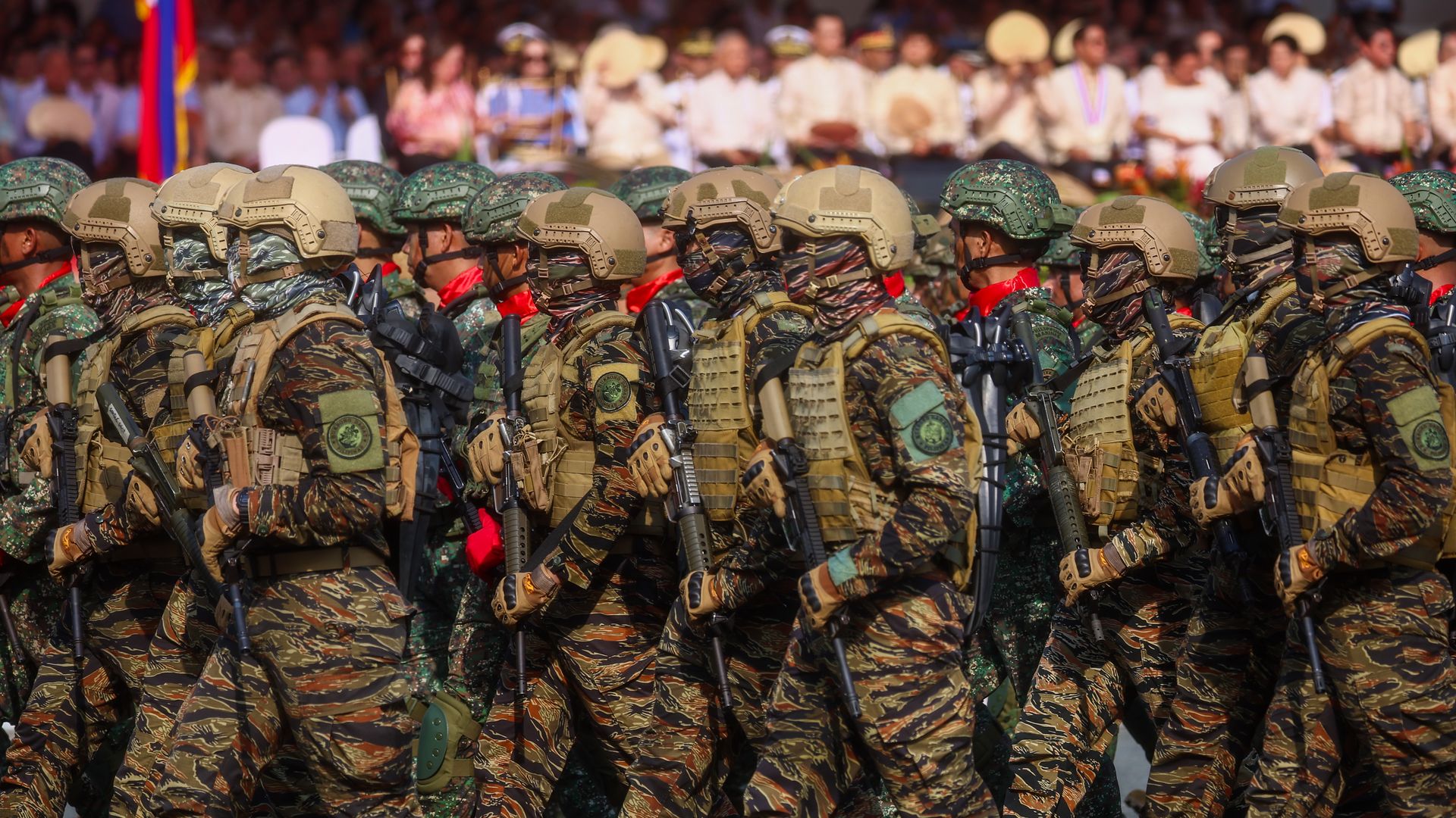 Rows of soldiers in camouflage uniforms and tan helmets march in formation, rifles at the ready. A blurred crowd and flag is seen behind them.