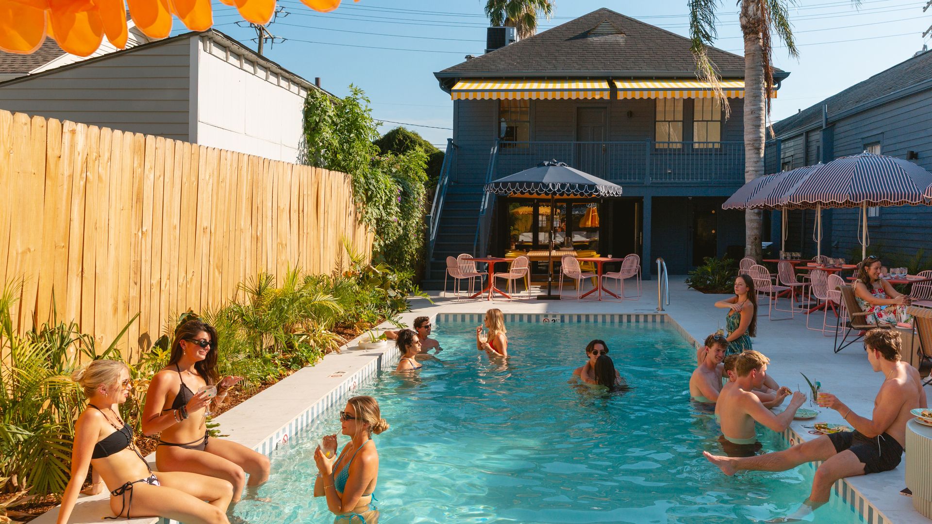 A sunny pool scene with people in swimsuits relaxing, chatting, and drinking by a blue pool surrounded by a wooden fence, plants, umbrellas, and a house with a yellow-striped awning.