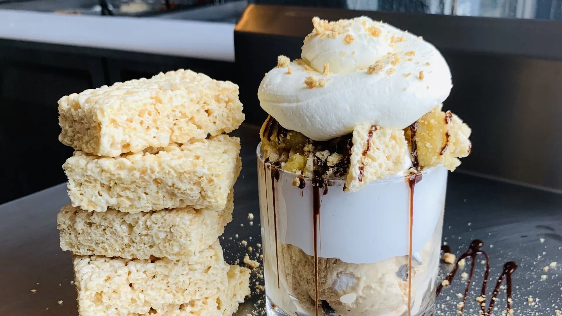 Stack of four light brown rice crispy treats next to a glass filled with dessert topped with whipped cream, rice crispy pieces, and chocolate drizzle.