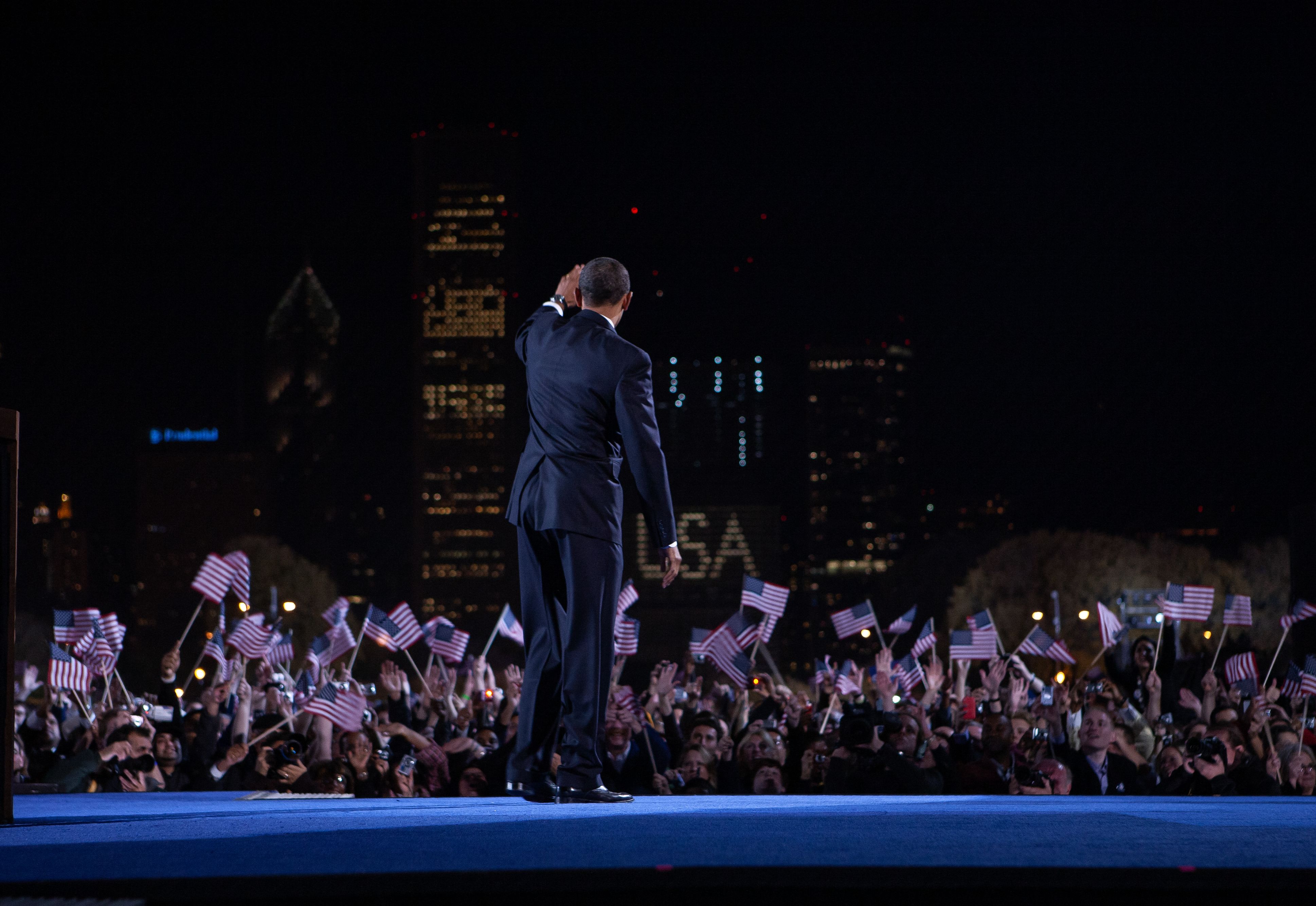 Nighttime outdoor rally with a man in a dark suit on stage, seen from behind, addressing a crowd waving numerous American flags; city skyline lights glow in the background.