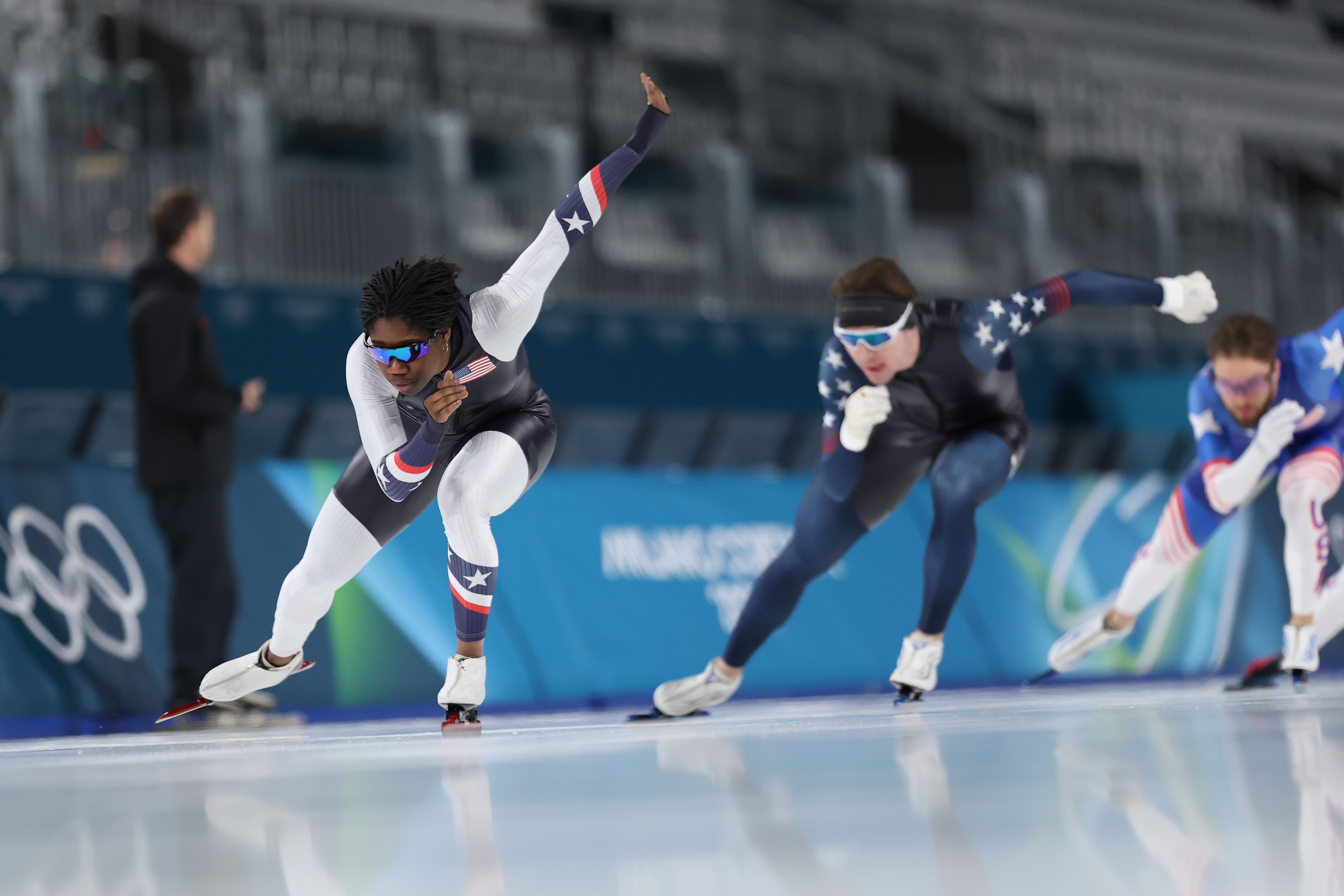 A woman speed-skates with two men speed skating behind her in an ice rink.