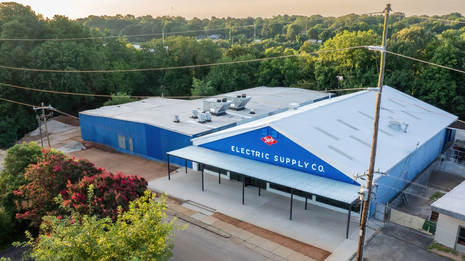 Blue warehouse building with "Electric Supply Co." sign in white letters, surrounded by green trees and pink flowering bushes, under a light sky at sunset.