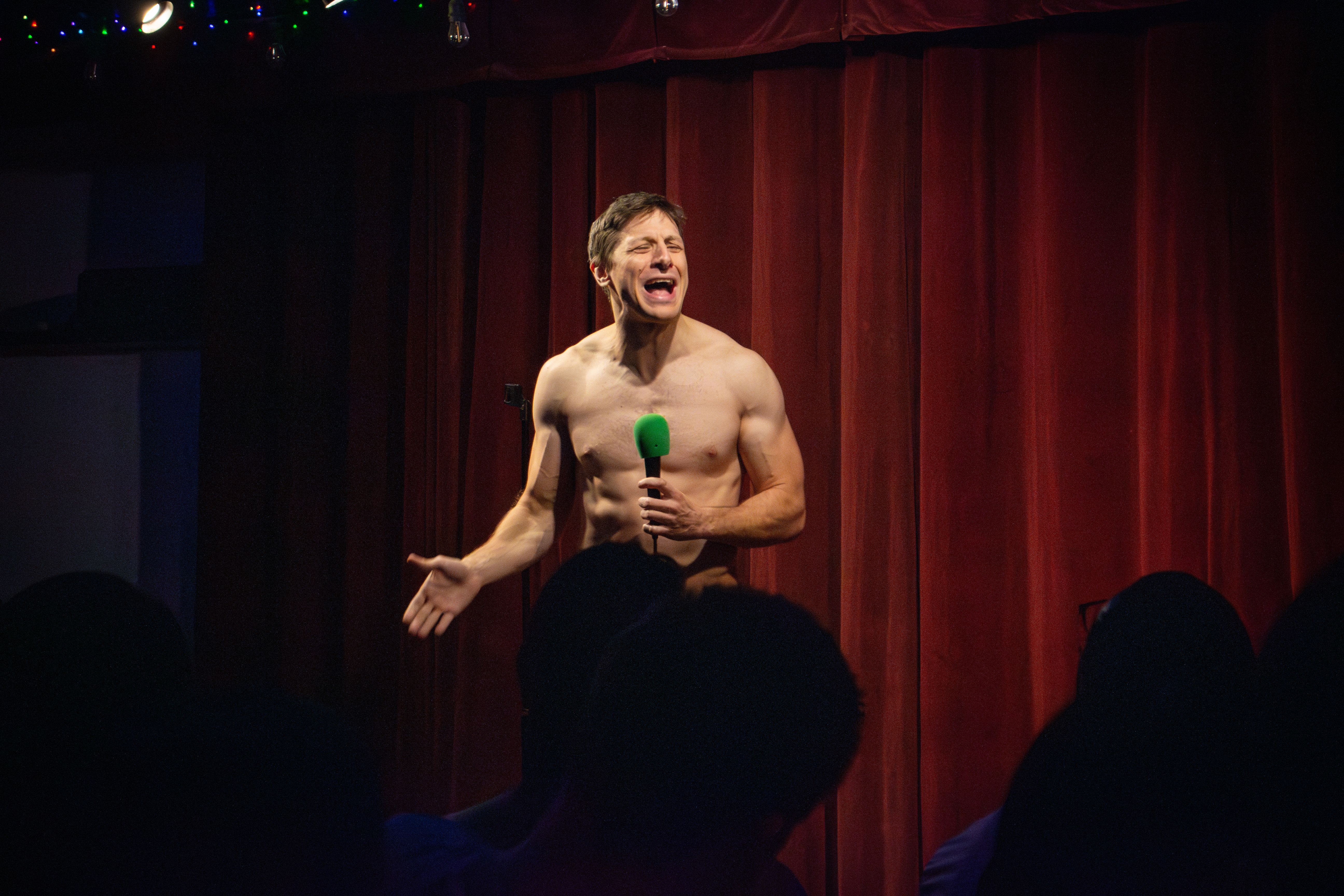 A shirtless male performer sings into a bright green microphone on a dark stage, with red theater curtains behind. Audience silhouettes fill the bottom, and colorful string lights sparkle above.