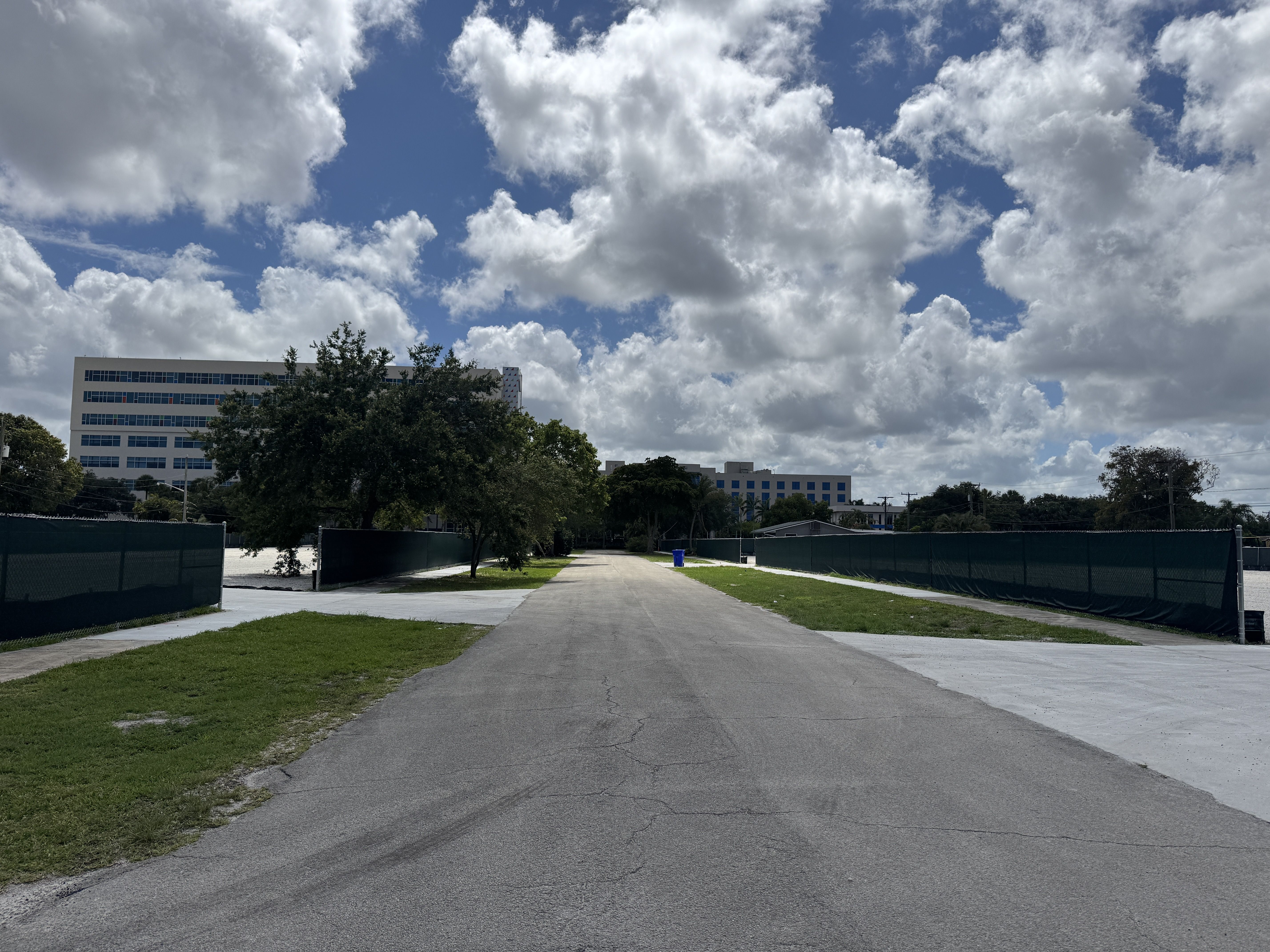 Wide street flanked by grass, trees, and fences, with a large white office building on the left and distant buildings beneath a bright blue sky with white clouds.