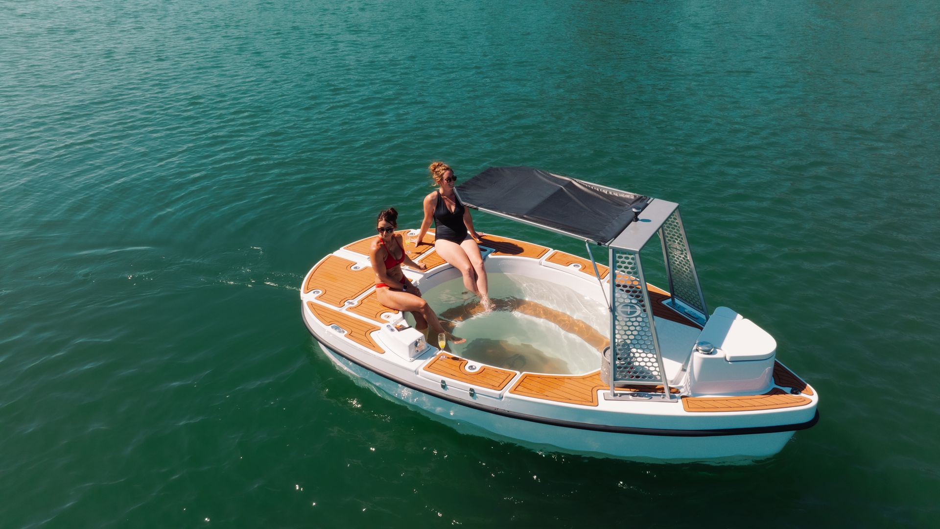 An aerial view of a white and orange pontoon boat with two women in swimsuits on the bow, turquoise water around, and a shaded canopy over the deck on a sunny day.