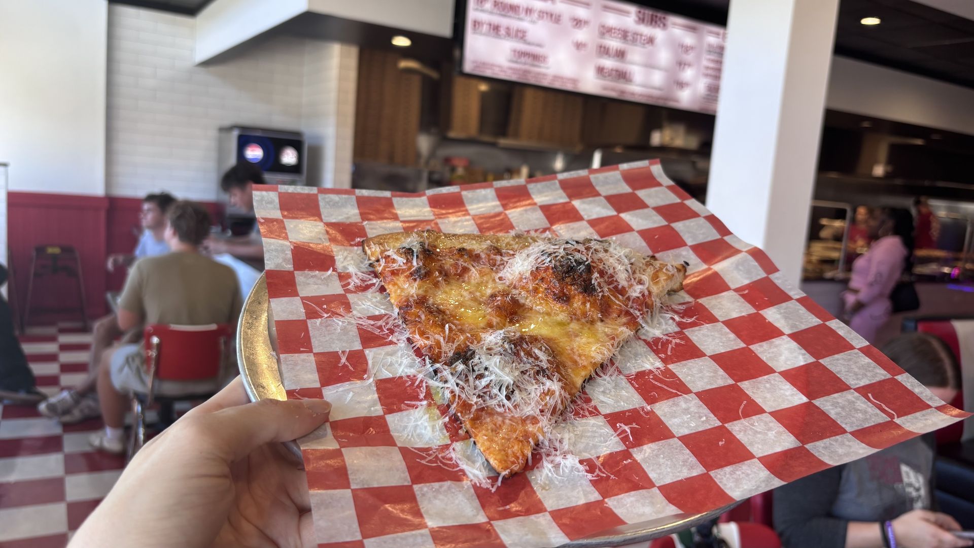A slice of cheesy pizza with stringy cheese on red-and-white checkered paper, held up in a casual pizza shop with blurred customers and a menu in the background.