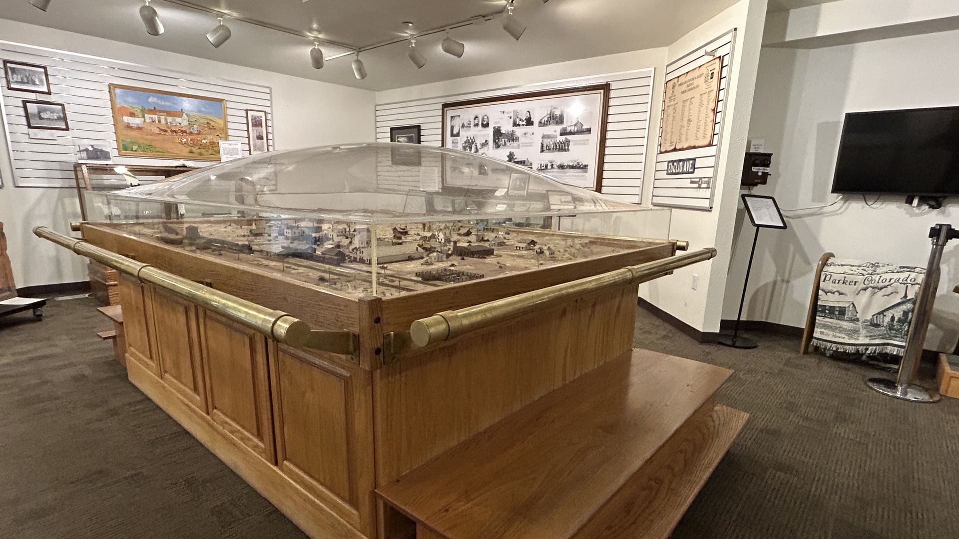 Museum diorama: a large wooden town model under glass on a raised base, with framed photos and panels around it; overhead track lighting and a banner about Parker, CO.
