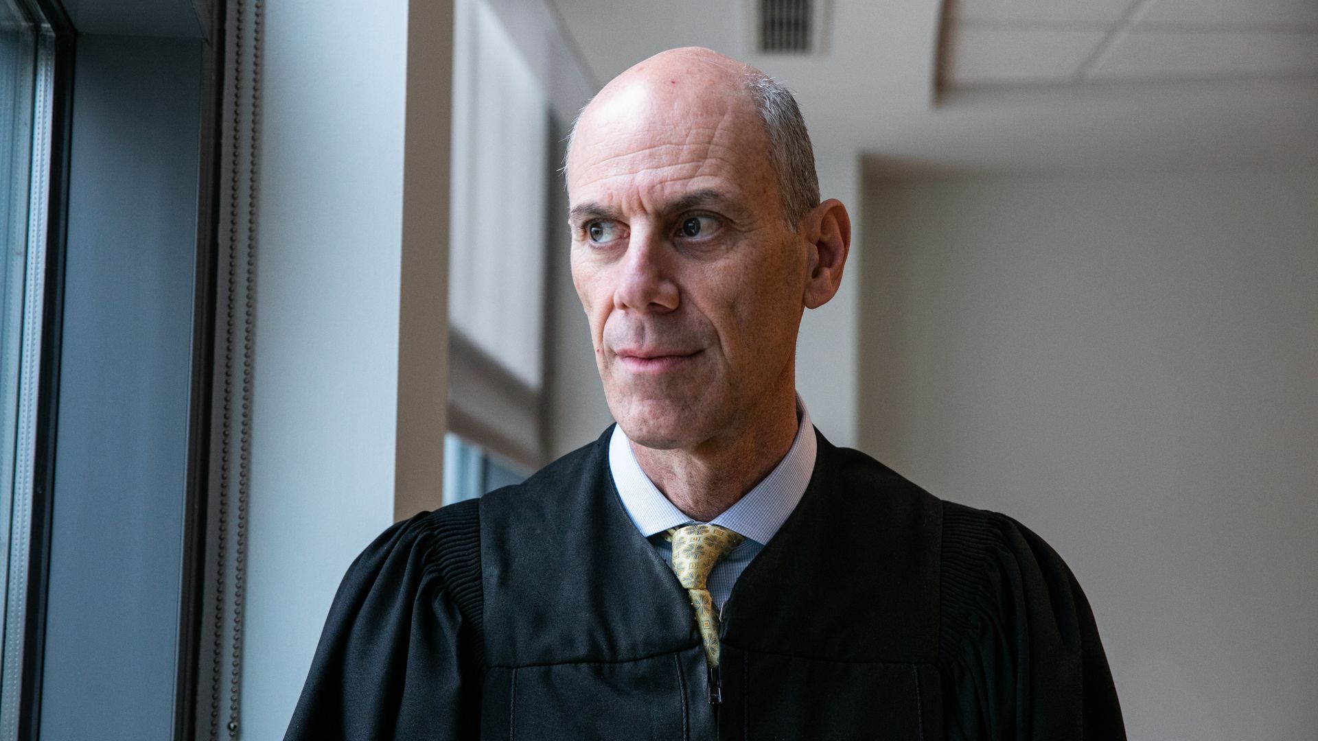 Judge Boasberg — wearing his black robe, a light-colored collared shirt and a yellow tie — looks out a window.