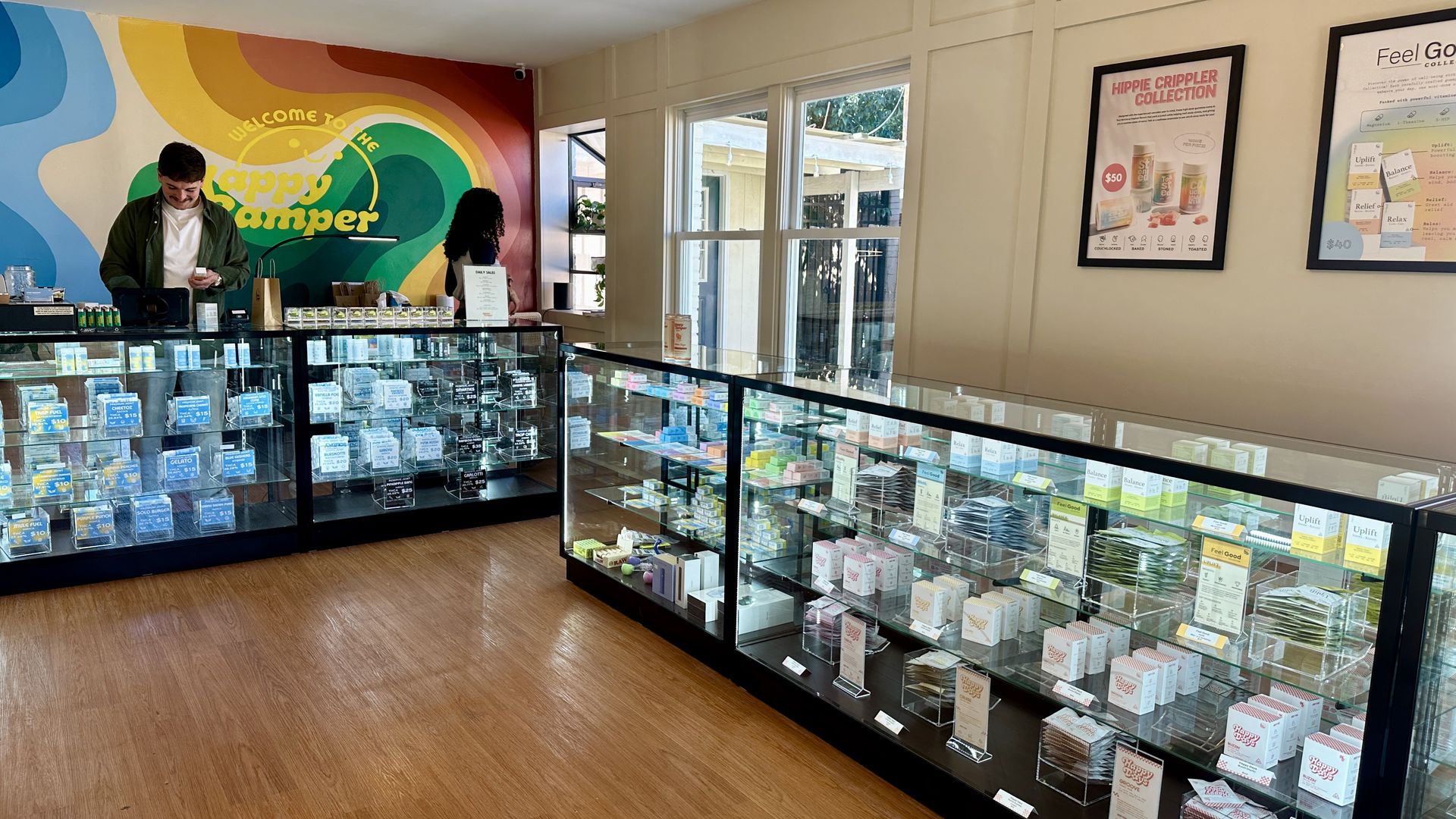 Bright shop interior with a rainbow mural reading Welcome to the Happy Camper behind glass display cases stocked with blue-labeled products; checkout counter on the left, window on the right.
