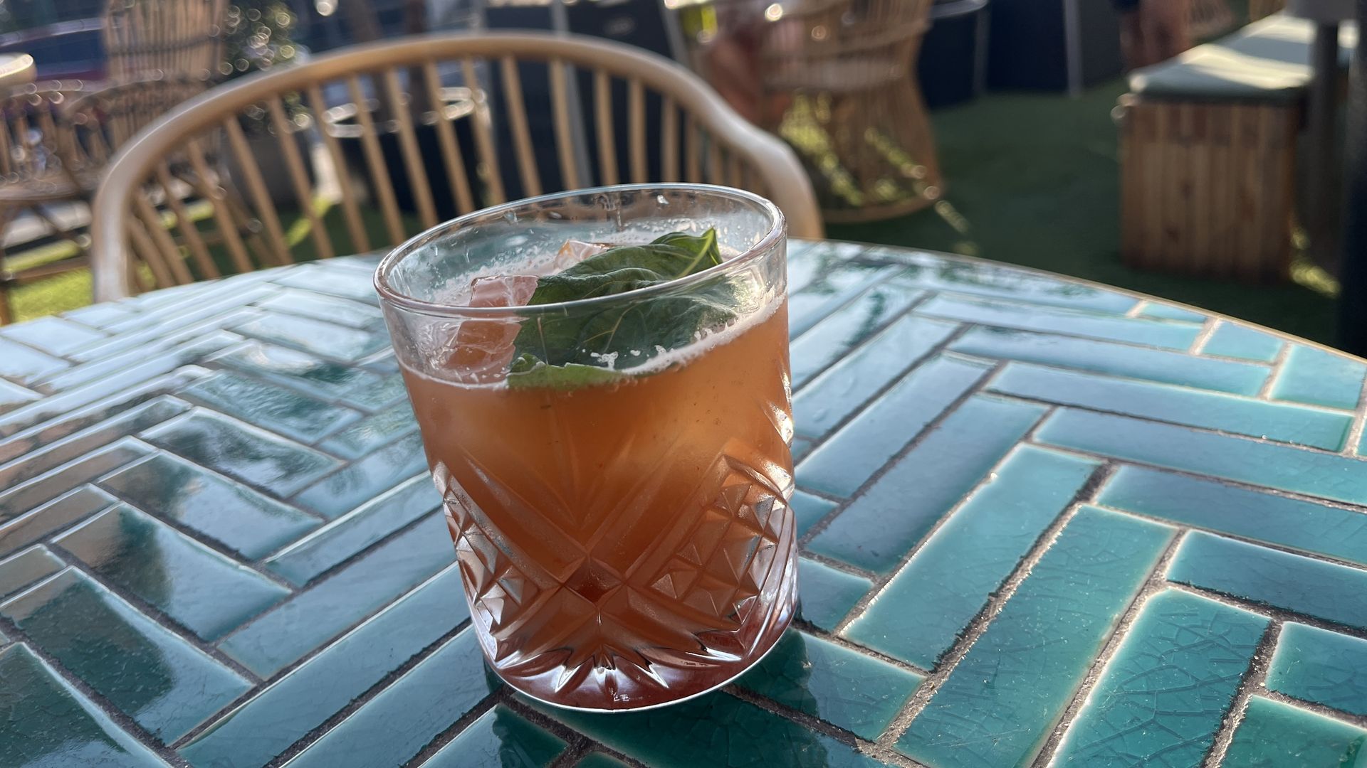 An orange drink in a cocktail glass topped with a basil leaf sits on a teal tiled table on an outdoor patio on a sunny day.