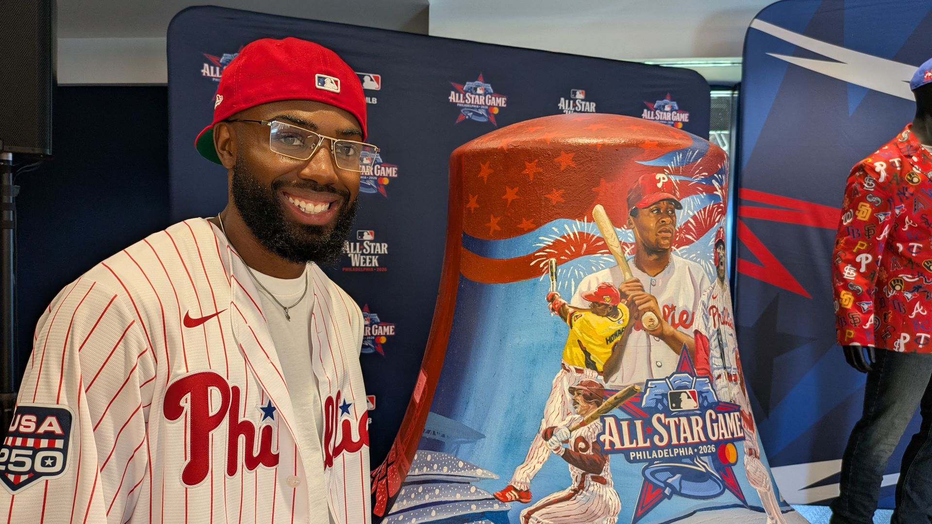 Smiling man in a white Phillies pinstripe jersey and red cap poses beside a large red All-Star Game display with stars and baseball imagery, against a Phillies backdrop.