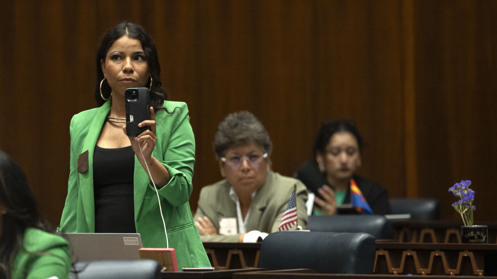 Analise Ortiz, wearing a green blazer over a black top, records video with her phone while standing at her desk on the floor of the Arizona House of Representatives, with several colleagues seated near her.
