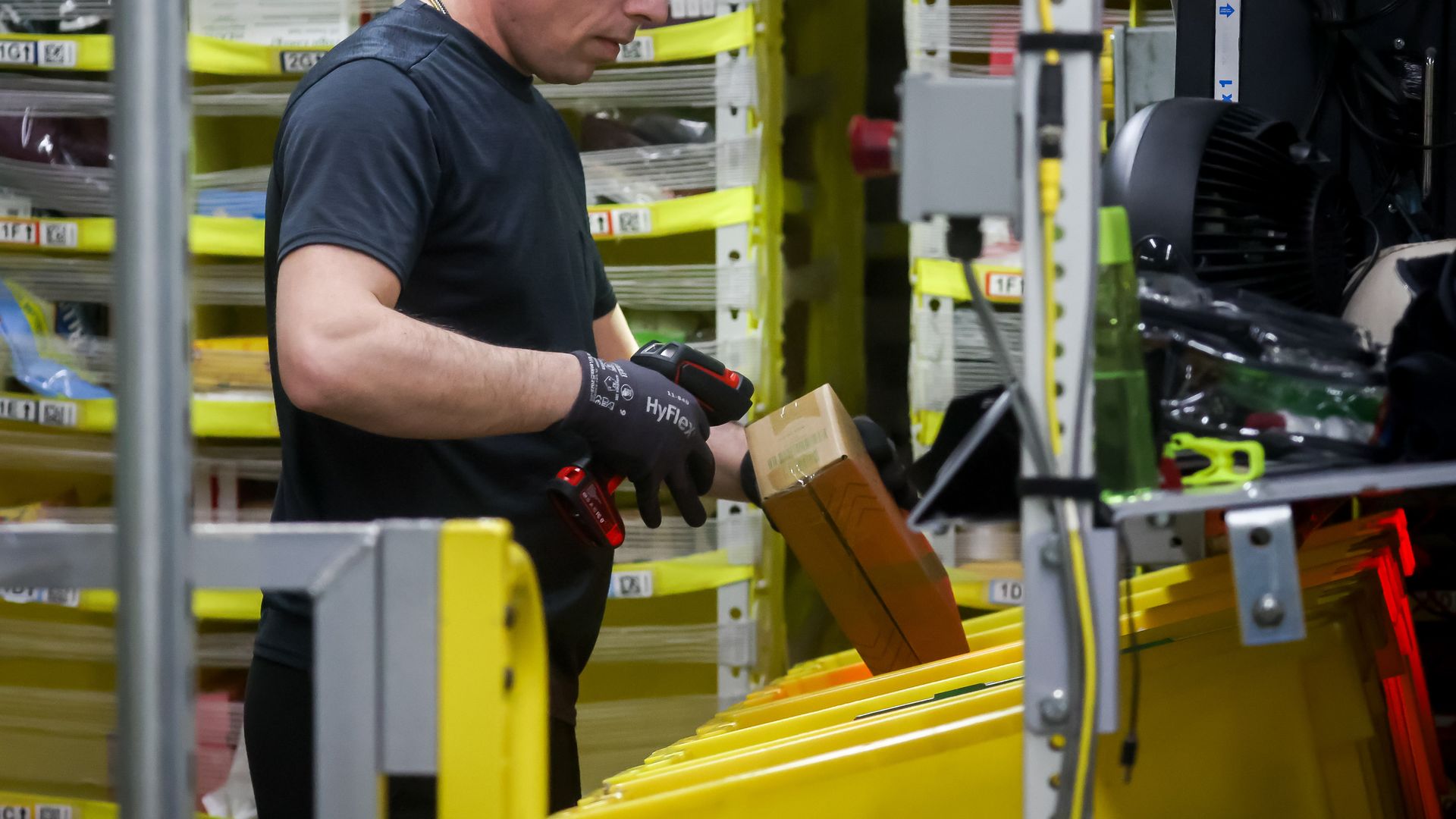 A warehouse worker in a dark T-shirt and HyFlex gloves handles a brown cardboard box with a red tool beside yellow storage racks and bins in an industrial warehouse.