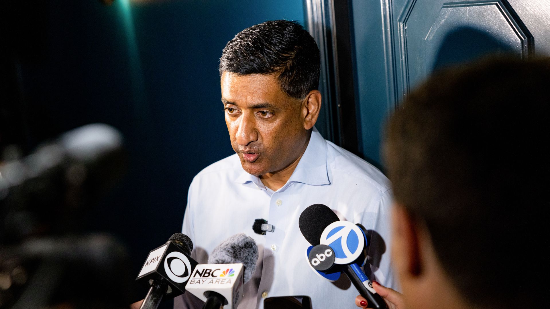 Man in a light blue shirt speaks to reporters; several microphones labeled ABC, NBC Bay Area, and KPIX are held up as a teal porta-potties appear in the background.
