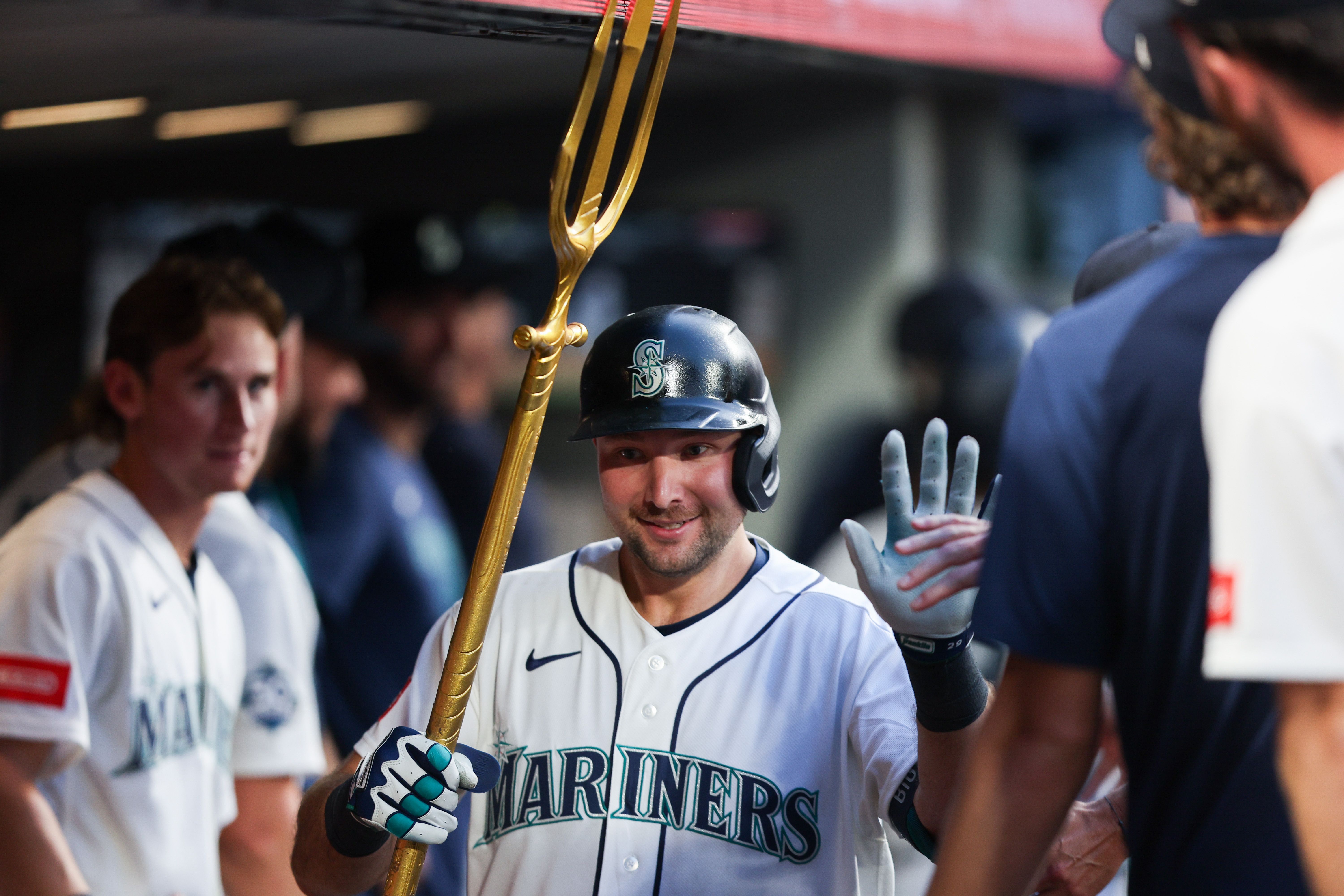 Cal Raleigh, wearing a Mariners jersey and batting helmet, holds a gold trident in one hand while holding up another hand, surrounded by other Mariners players and support staff.